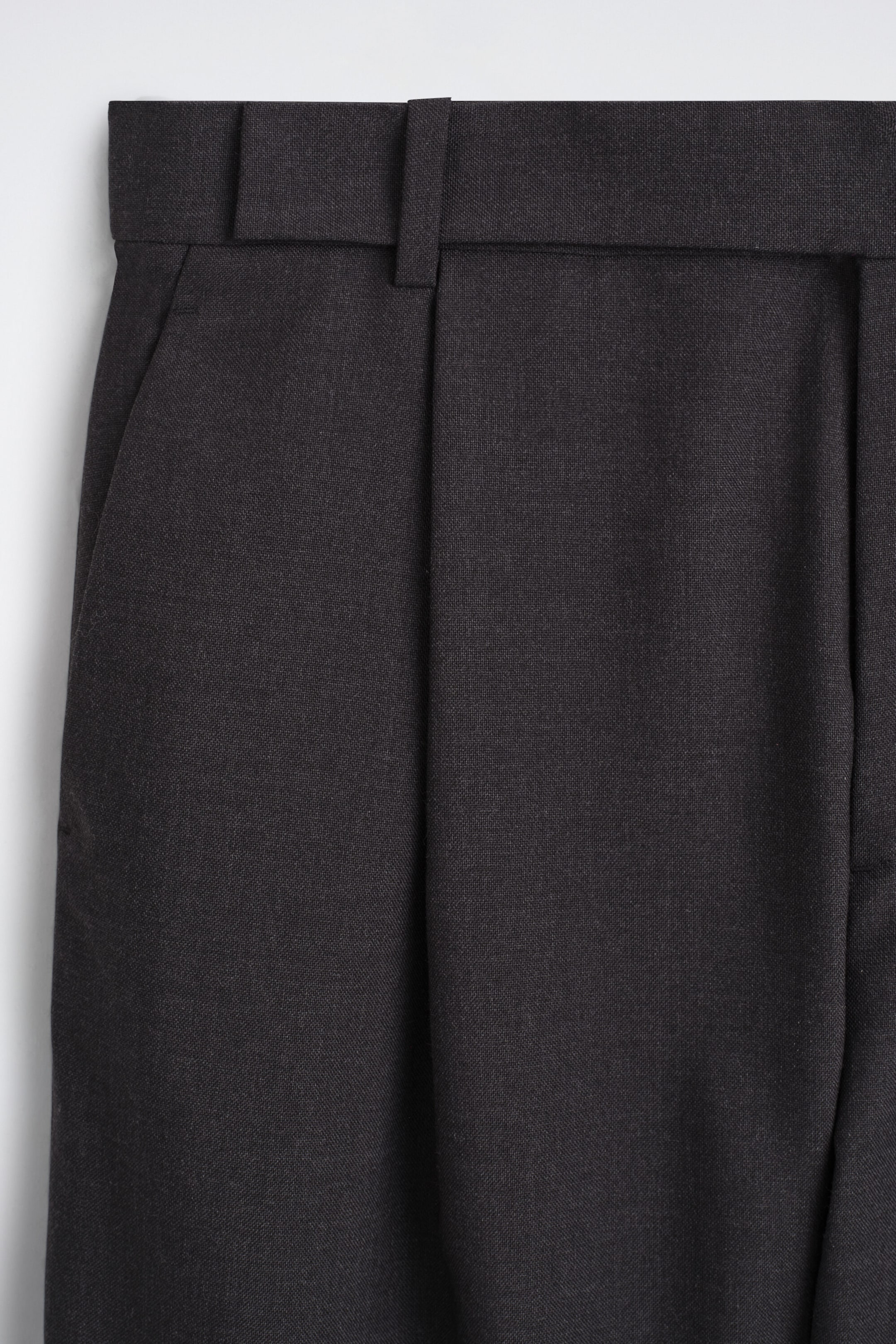 These dark grey wool relaxed-fit barrel-leg trousers feature a close-up front view of the structured waistband, integrated belt loops, and a side slip pocket. A sharp vertical pleat runs down from the waistline, highlighting the tailored construction of the textured fabric. These dark grey wool relaxed-fit barrel-leg trousers feature a close-up front view of the structured waistband, integrated belt loops, and a side slip pocket. A sharp vertical pleat runs down from the waistline, highlighting the tailored construction of the textured fabric.
