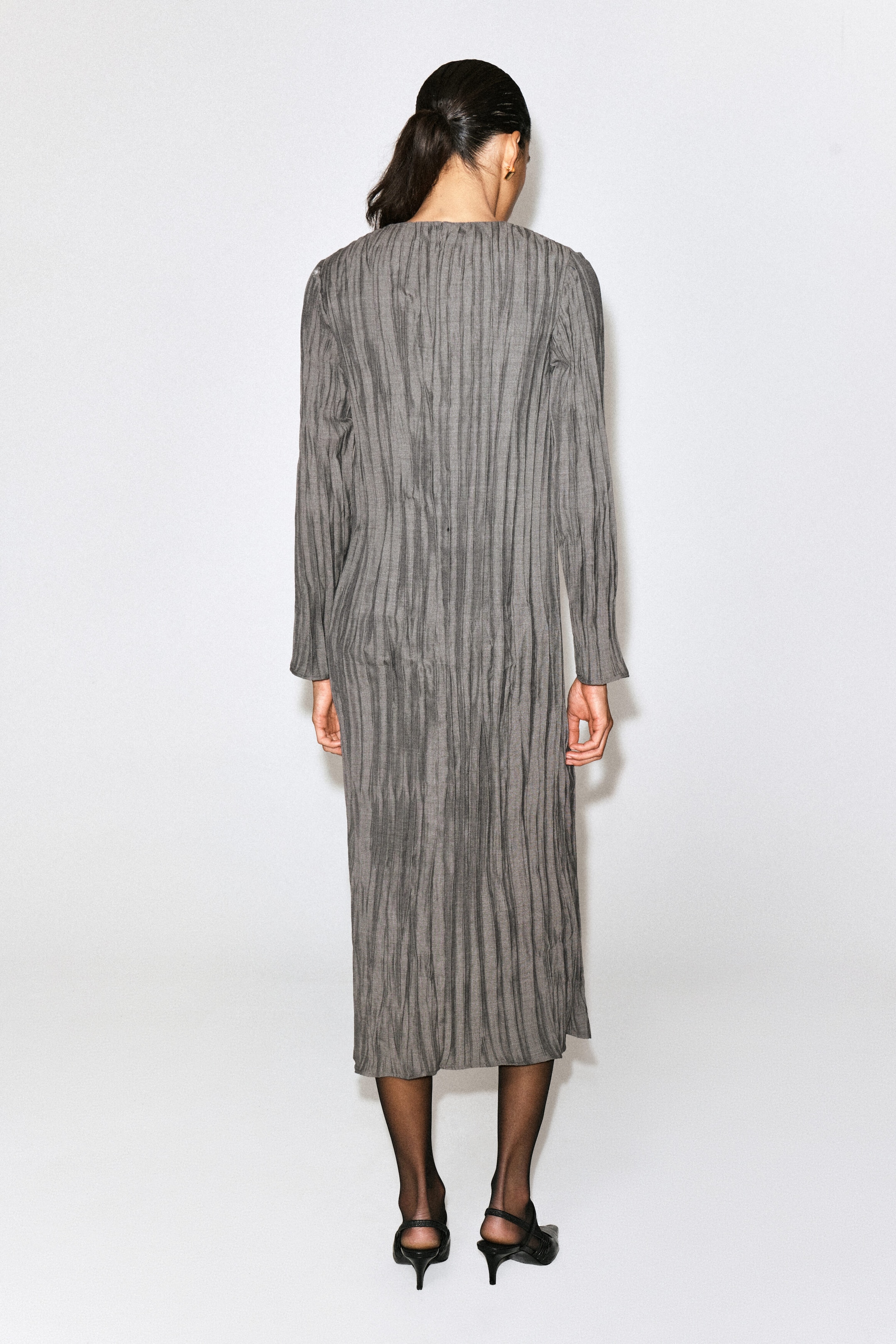 A gray long-sleeved midi dress is shown from the back, highlighting its straight silhouette and all-over vertical crinkle-pleated texture. The dress drapes loosely to mid-calf, emphasizing its relaxed structure and uniform texture.