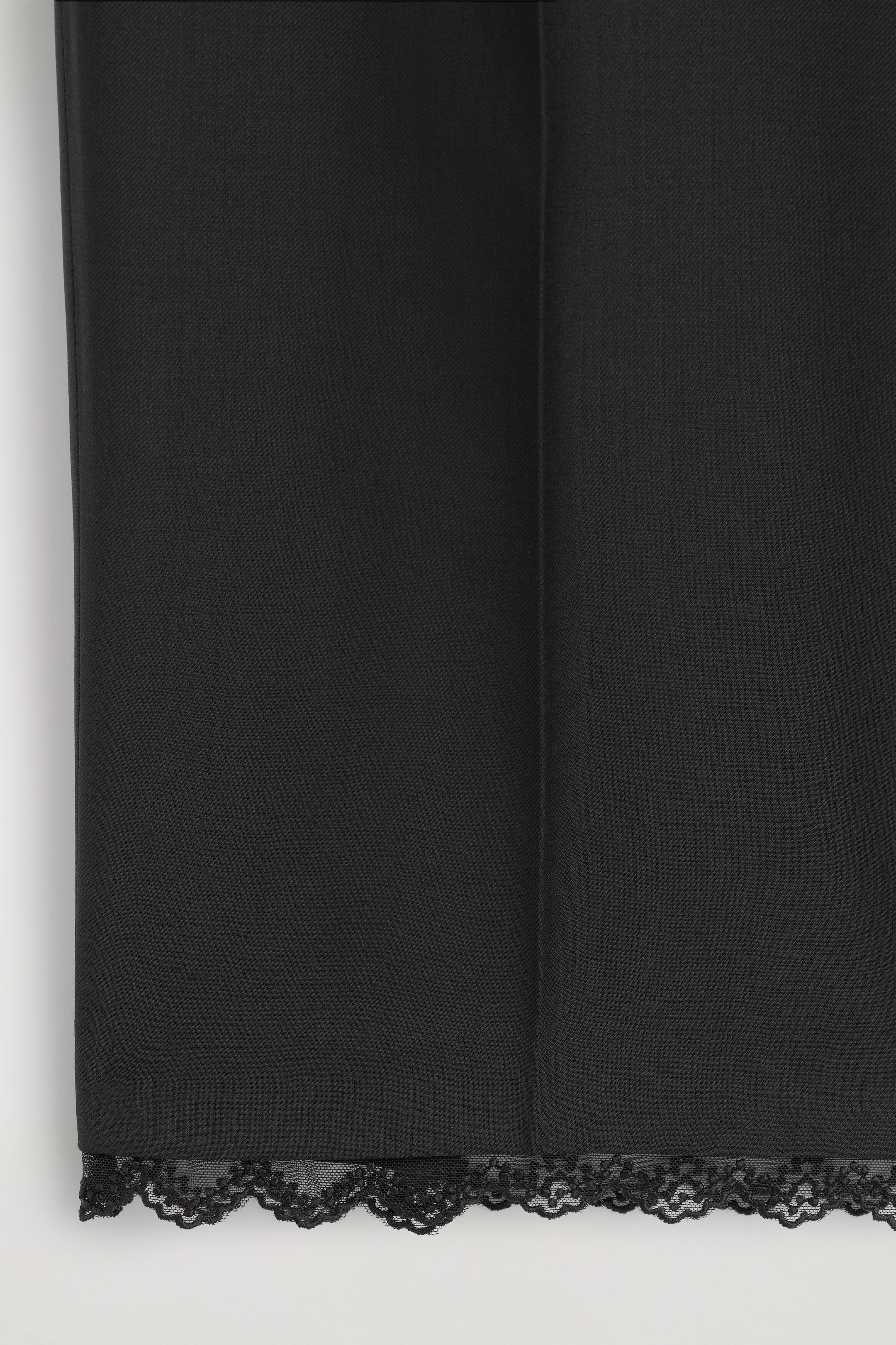 A close-up of the front of black trousers, showcasing a sharp vertical pressed crease and a delicate black scalloped lace trim along the bottom hem. A close-up of the front of black trousers, showcasing a sharp vertical pressed crease and a delicate black scalloped lace trim along the bottom hem.