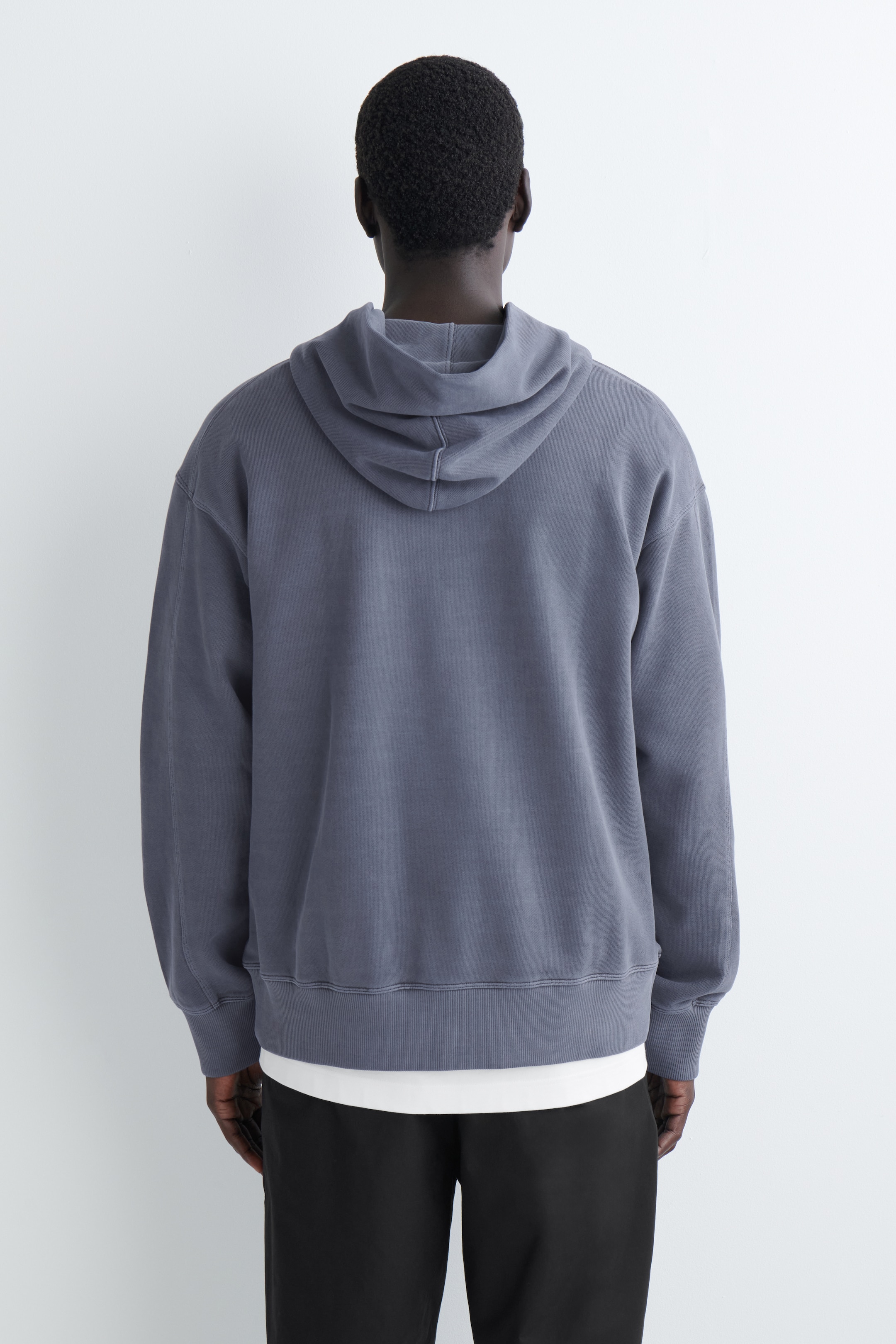 Grösseres Bild ansehen: Ein gewaschener hoodie in Anthrazitgrau wird aus der Rückansicht präsentiert, mit einem entspannten Schnitt, langen Ärmeln mit gerippten Bündchen und einem gerippten Saum. Ein weißes T-Shirt ist sichtbar darunter geschichtet, das unterhalb des Saums des Hoodies hervorsteht.