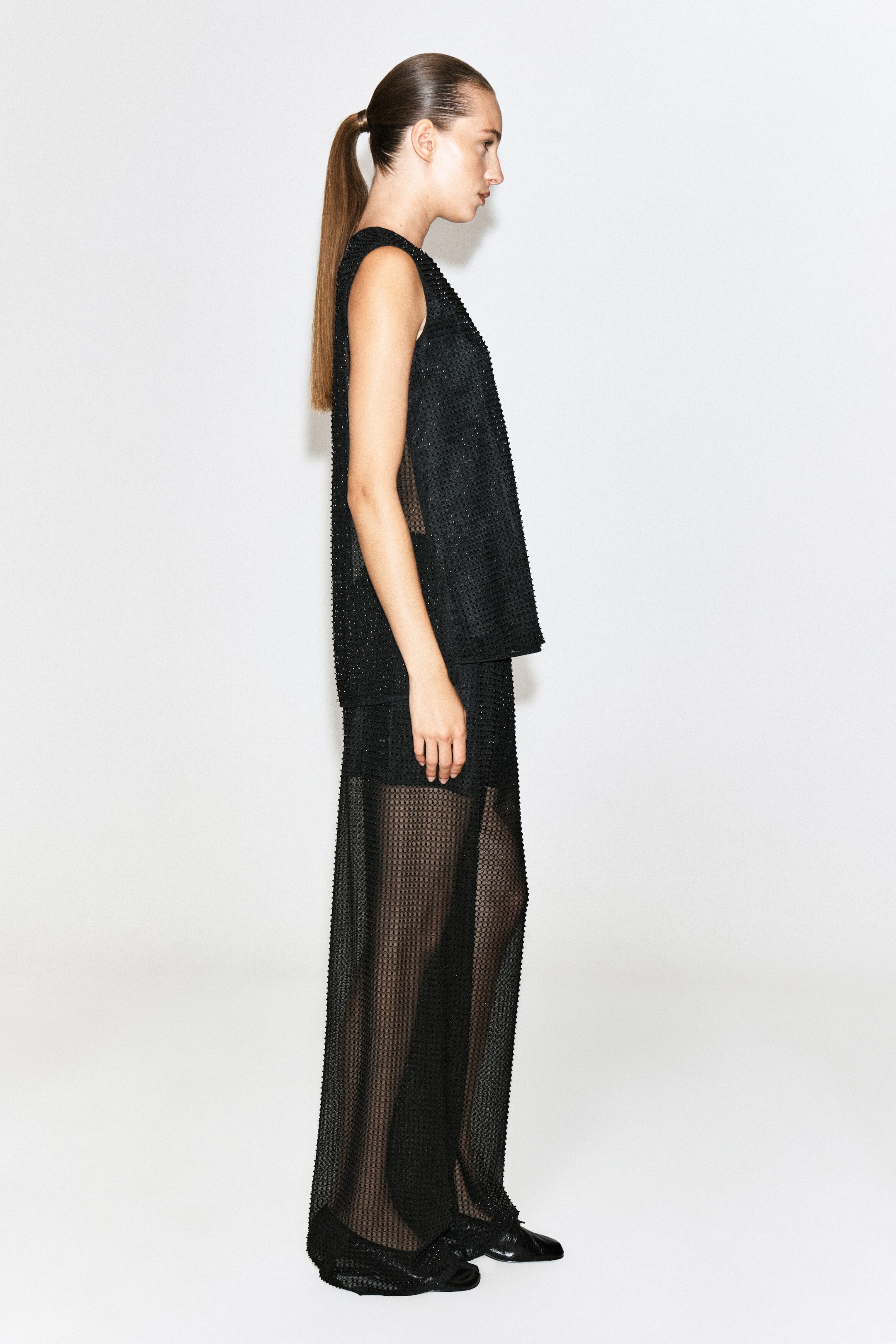 Beaded knitted trousers in a sheer black open-knit fabric are being worn by the model standing in profile. The wide-leg trousers feature subtle all-over bead embellishments and are styled with a matching sleeveless top.