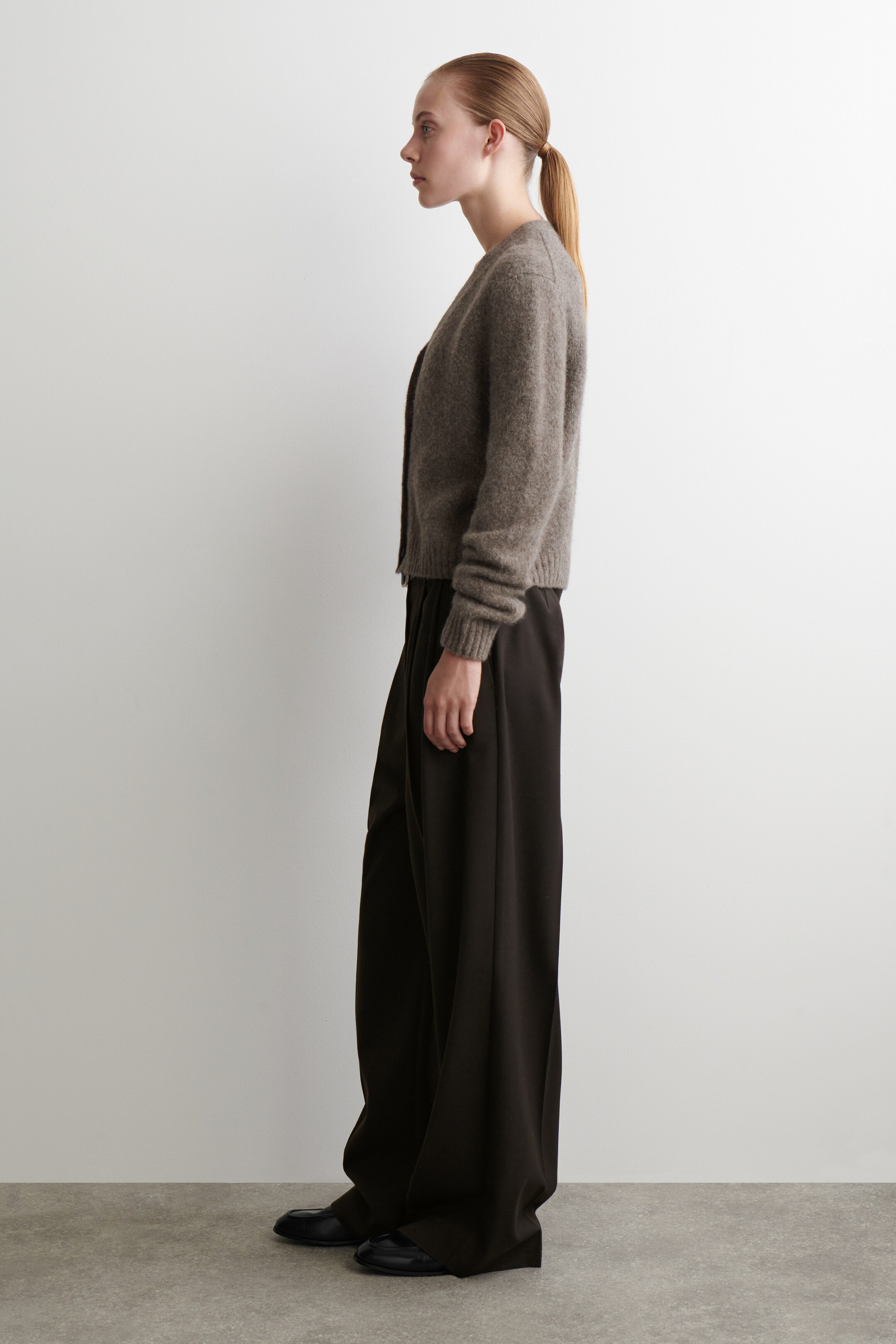 Se større billede: En dæmpet brun, fuzzy strikket cardigan præsenteres i en sideprofil, med en V-hals, knaplukning foran og ribbede manchetter og kant. Den er parret med mørke, brede bukser, der samler sig ved anklerne, og sorte loafers bæres nedenunder.