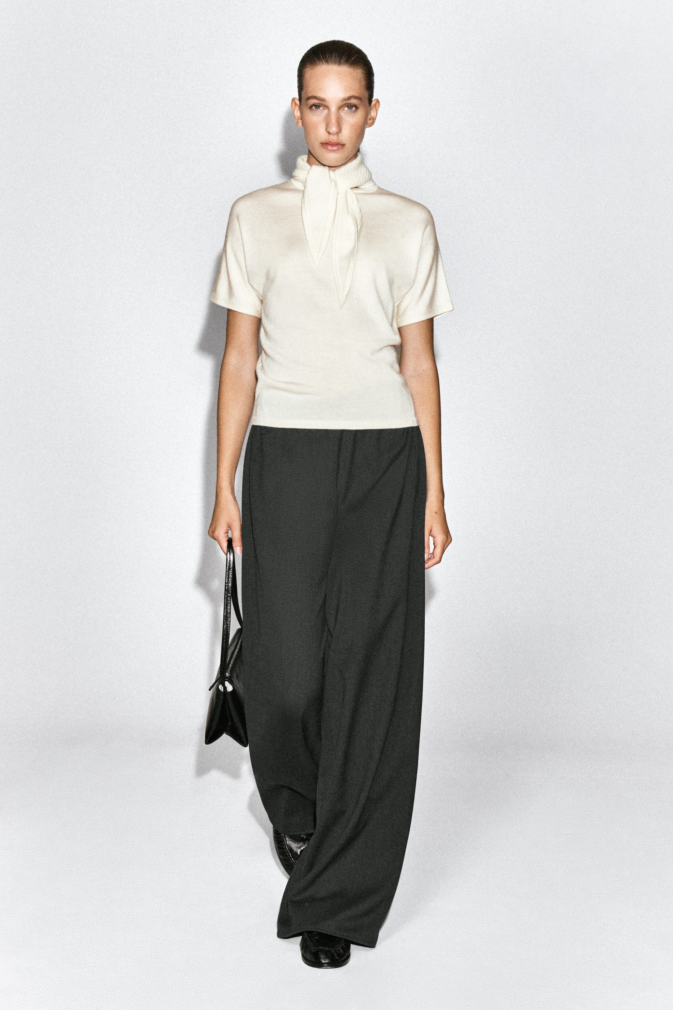The cream scarf-detail merino wool-blend T-shirt is worn by the model and features short sleeves and an integrated scarf collar tied at the front of the neck. It is styled with dark wide-leg trousers, and a black handbag is being carried in one hand.