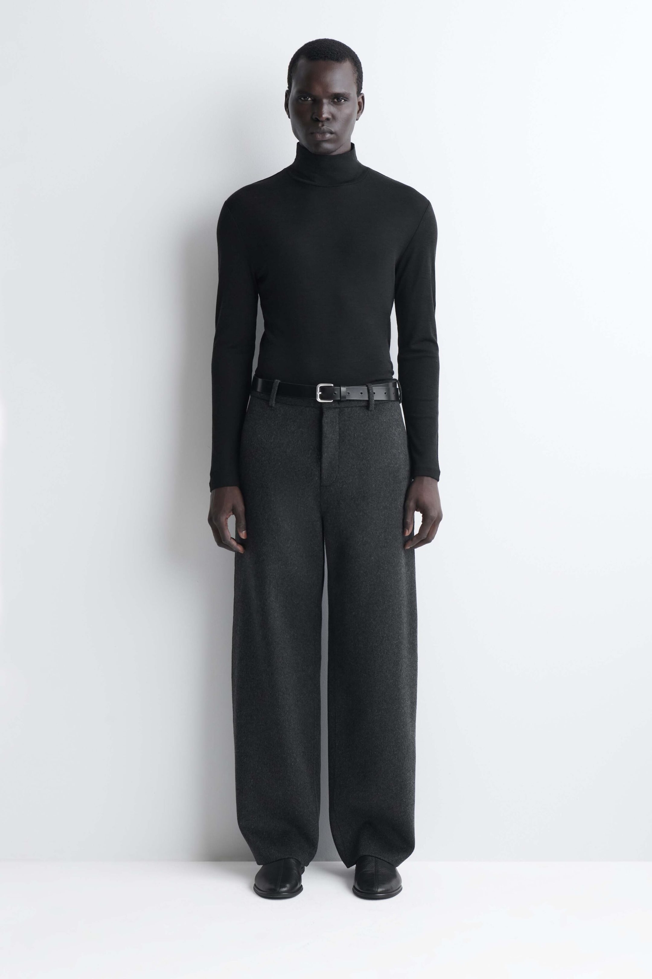 The slim wool jersey roll neck top in black is shown from the front, featuring a close-fitting silhouette, a high folded collar, and long sleeves. The top is worn tucked into dark grey trousers with a black belt, highlighting its streamlined, lightweight knit construction. The slim wool jersey roll neck top in black is shown from the front, featuring a close-fitting silhouette, a high folded collar, and long sleeves. The top is worn tucked into dark grey trousers with a black belt, highlighting its streamlined, lightweight knit construction.
