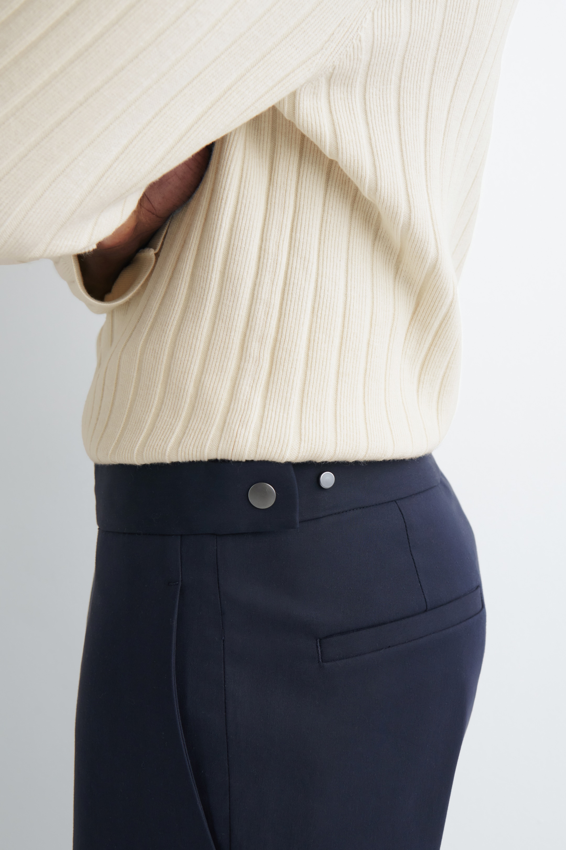 Vezi imaginea mai mare: Un pulover crem cu nervuri, cu nervuri verticale vizibile, este purtat și este băgat în pantaloni bleumarin. Pantalonii au o talie lată cu o închidere asimetrică, care este asigurată de un capse mari și unul mai mic de culoare argintie. Un buzunar lateral și un buzunar cu refilet la spate sunt, de asemenea, vizibile pe șolduri.