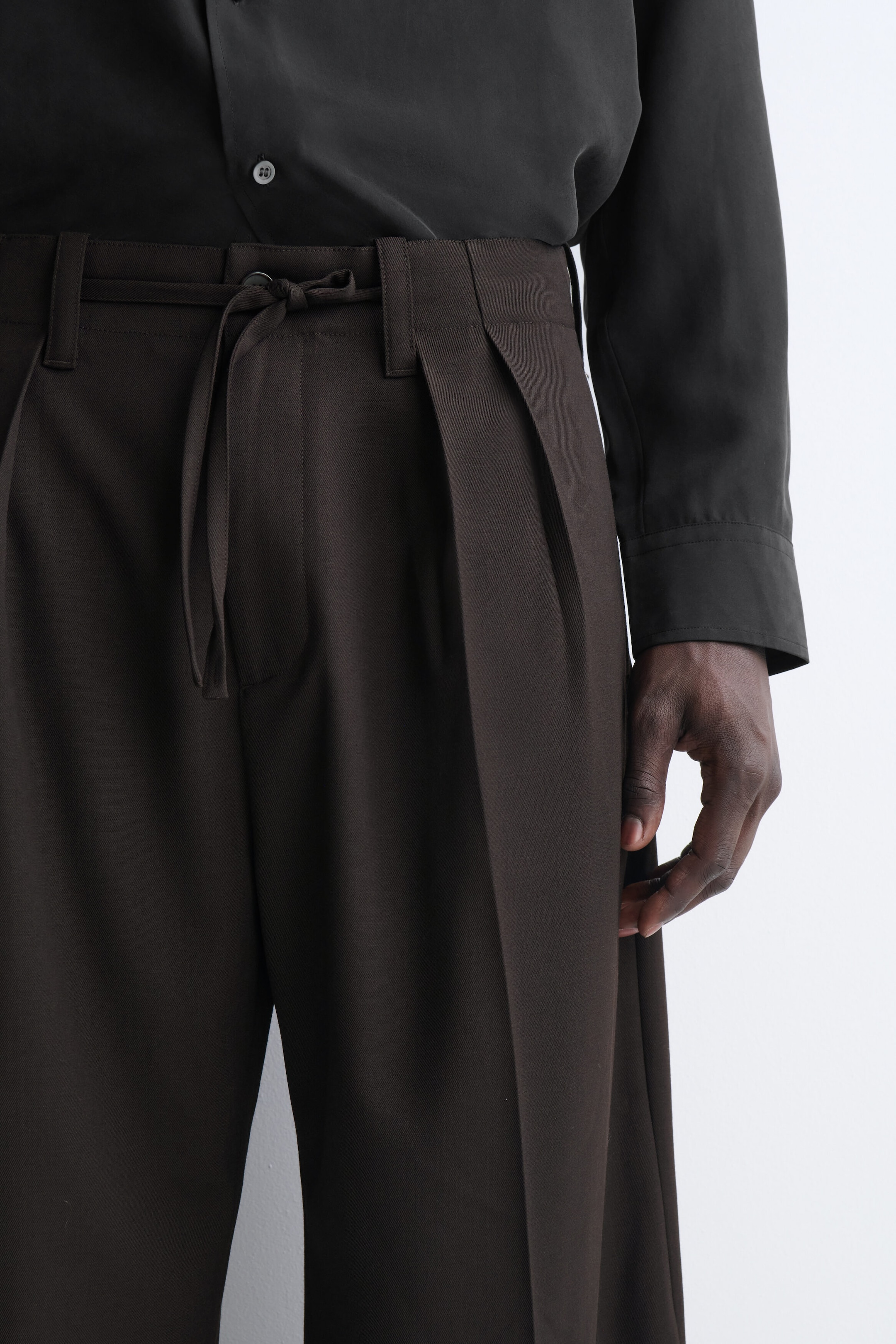 A close-up of the top portion of the dark brown trousers shows a waistband with belt loops, a matching fabric drawstring tie, and deep front pleats. The trousers are styled with a tucked-in black long-sleeve button-up shirt, highlighting their structured fit. A close-up of the top portion of the dark brown trousers shows a waistband with belt loops, a matching fabric drawstring tie, and deep front pleats. The trousers are styled with a tucked-in black long-sleeve button-up shirt, highlighting their structured fit.