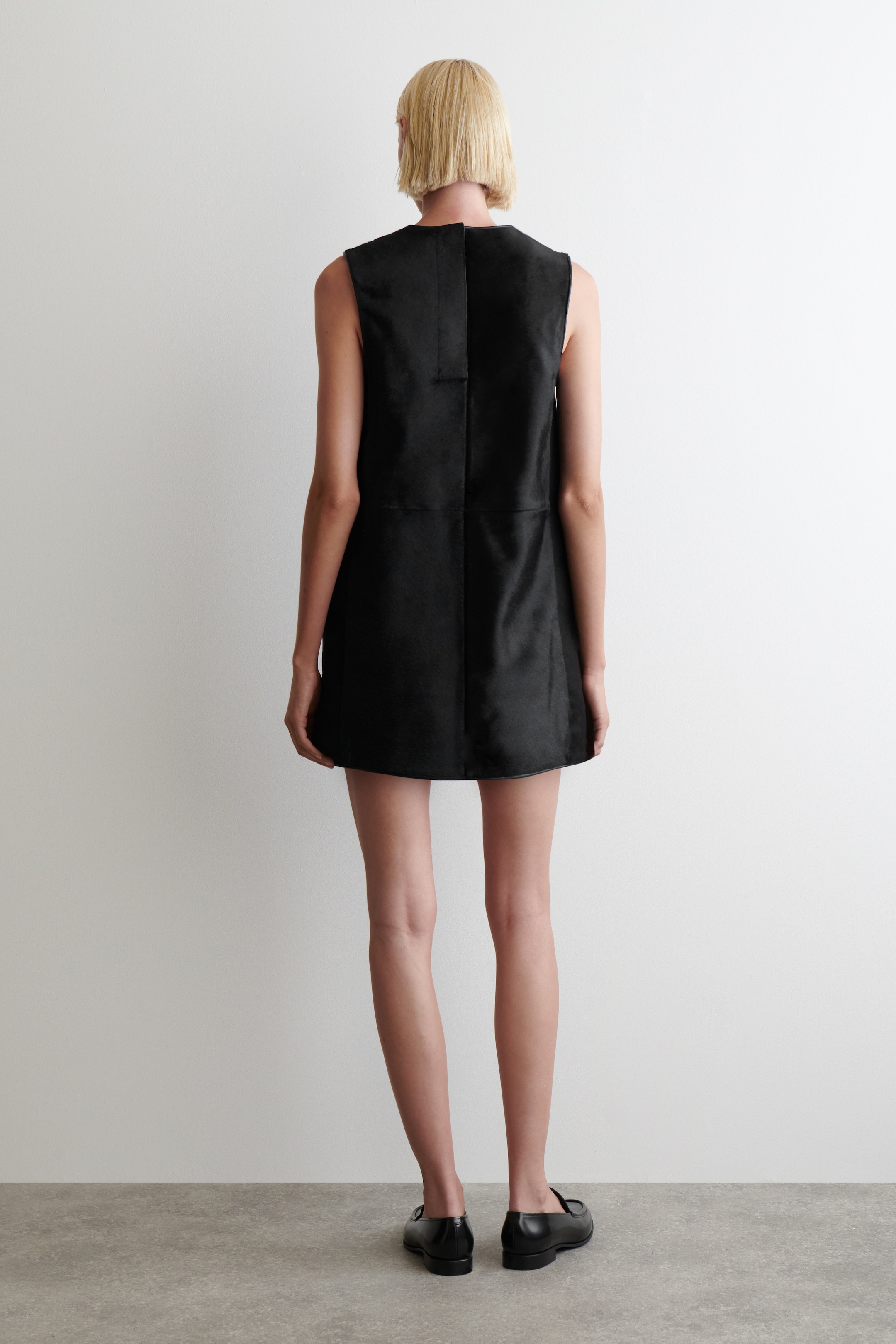 Agrandir l'image: Une robe noire sans manches est présentée de dos, avec une silhouette évasée en A et une longueur mini. Des coutures verticales et horizontales proéminentes détaillent l'arrière de son tissu à finition mate.