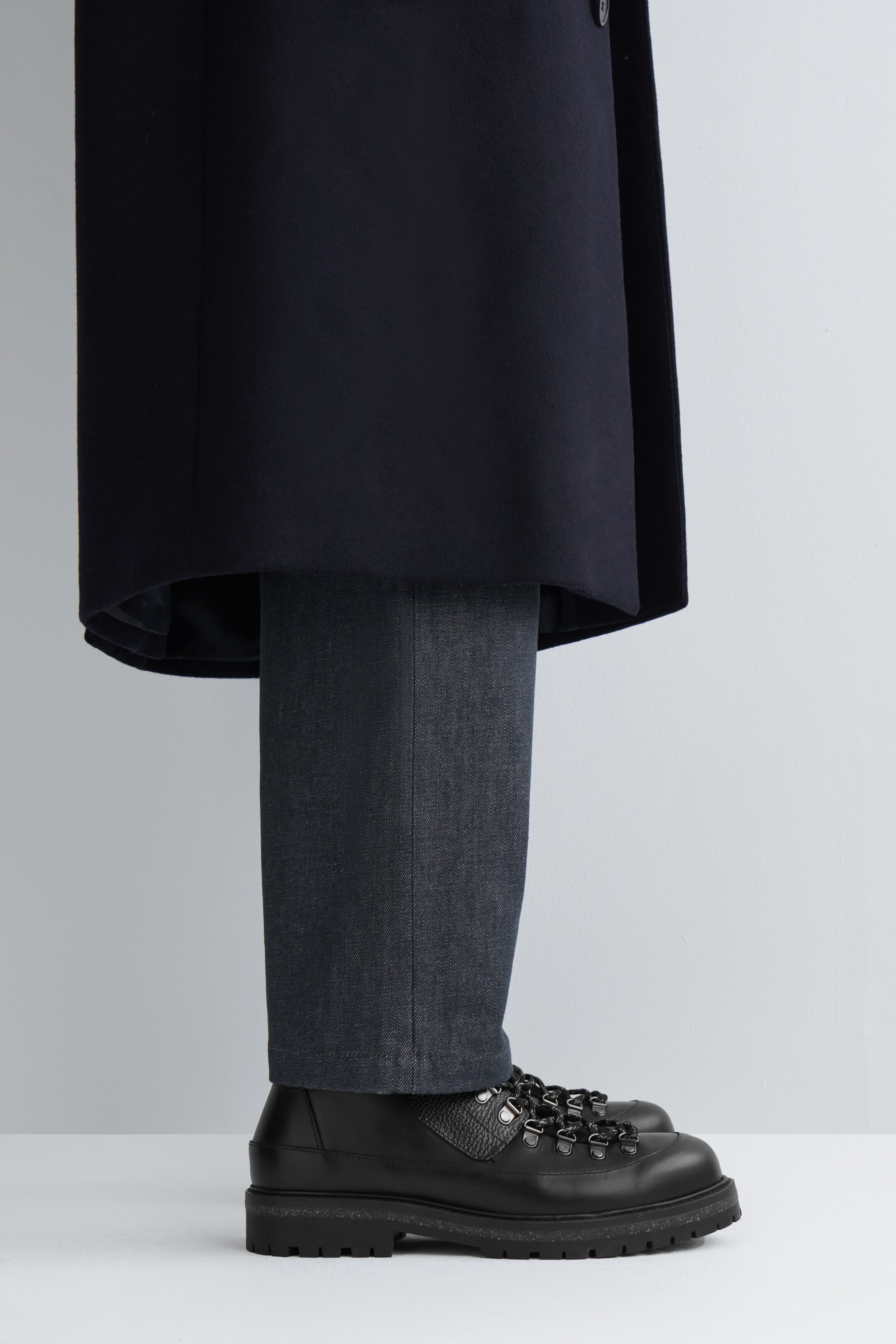 Visualizza immagine più grande: Un cappotto blu scuro è stratificato su pantaloni grigio scuro e testurizzati, che si estendono fino a un paio di scarponi da trekking neri. Gli scarponi presentano occhielli in metallo a forma di D, un dettaglio in pelle martellata sulla parte superiore e una suola spessa e dentellata, con il loro profilo rivolto in avanti.