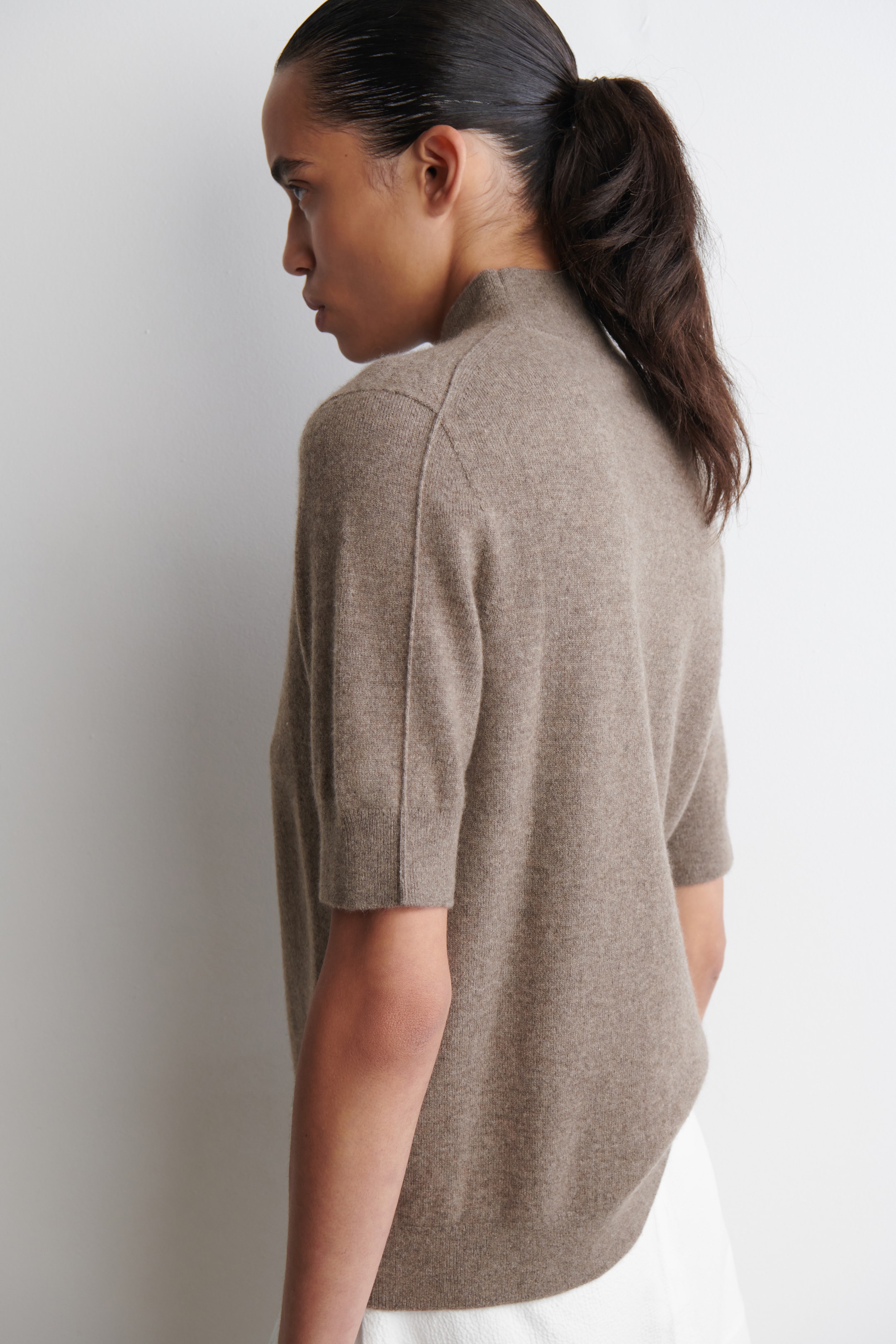 Agrandir l'image: Un pull en tricot brun est présenté de dos à 3/4, mettant en valeur ses manches courtes et son col montant. Des détails de couture visibles longent les épaules et descendent le long des côtés, tandis que l'ourlet du pull se courbe sur un vêtement blanc visible.