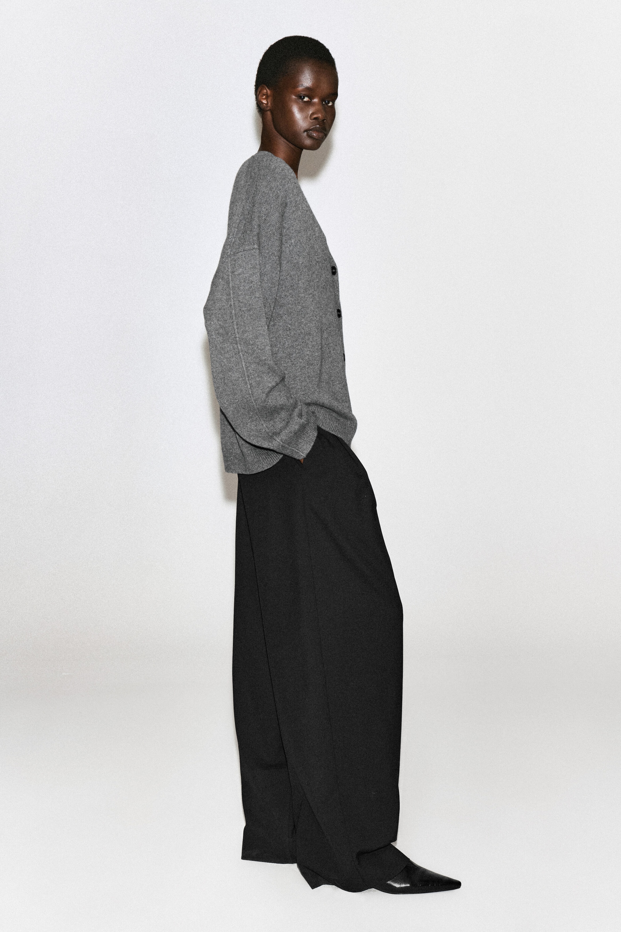 La modella indossa un cardigan grigio in cashmere con scollo a V, caratterizzato da una vestibilità rilassata, spalle scese e bottoni frontali scuri. Il cardigan è abbinato a pantaloni neri a gamba larga e scarpe nere con punta affusolata, mentre la modella posa di profilo laterale. La modella indossa un cardigan grigio in cashmere con scollo a V, caratterizzato da una vestibilità rilassata, spalle scese e bottoni frontali scuri. Il cardigan è abbinato a pantaloni neri a gamba larga e scarpe nere con punta affusolata, mentre la modella posa di profilo laterale.