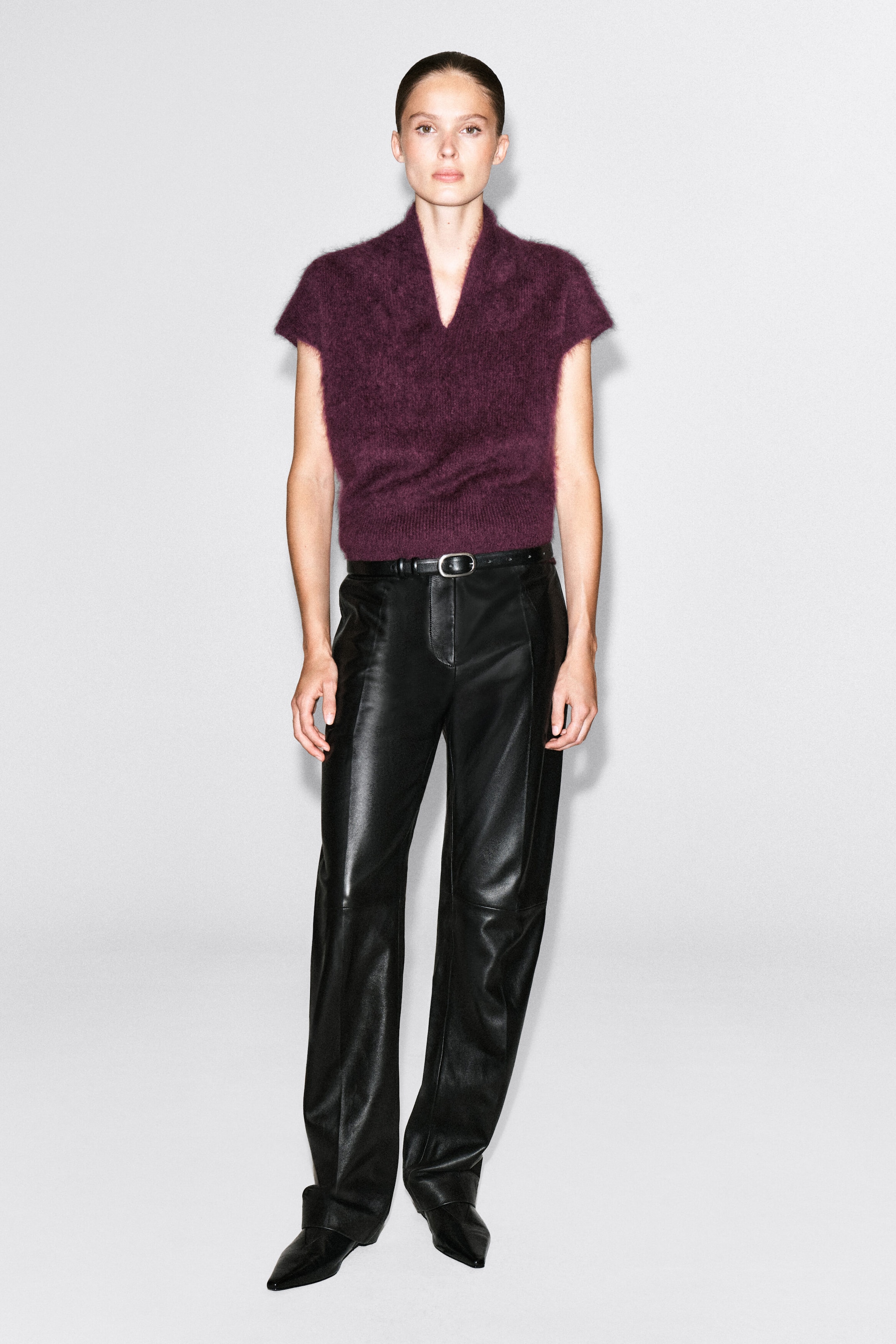 A draped, brushed merino wool top in a deep plum color is worn by the model, featuring a V-neckline, short sleeves, and a fuzzy texture. The top is tucked into black leather trousers styled with a black belt.