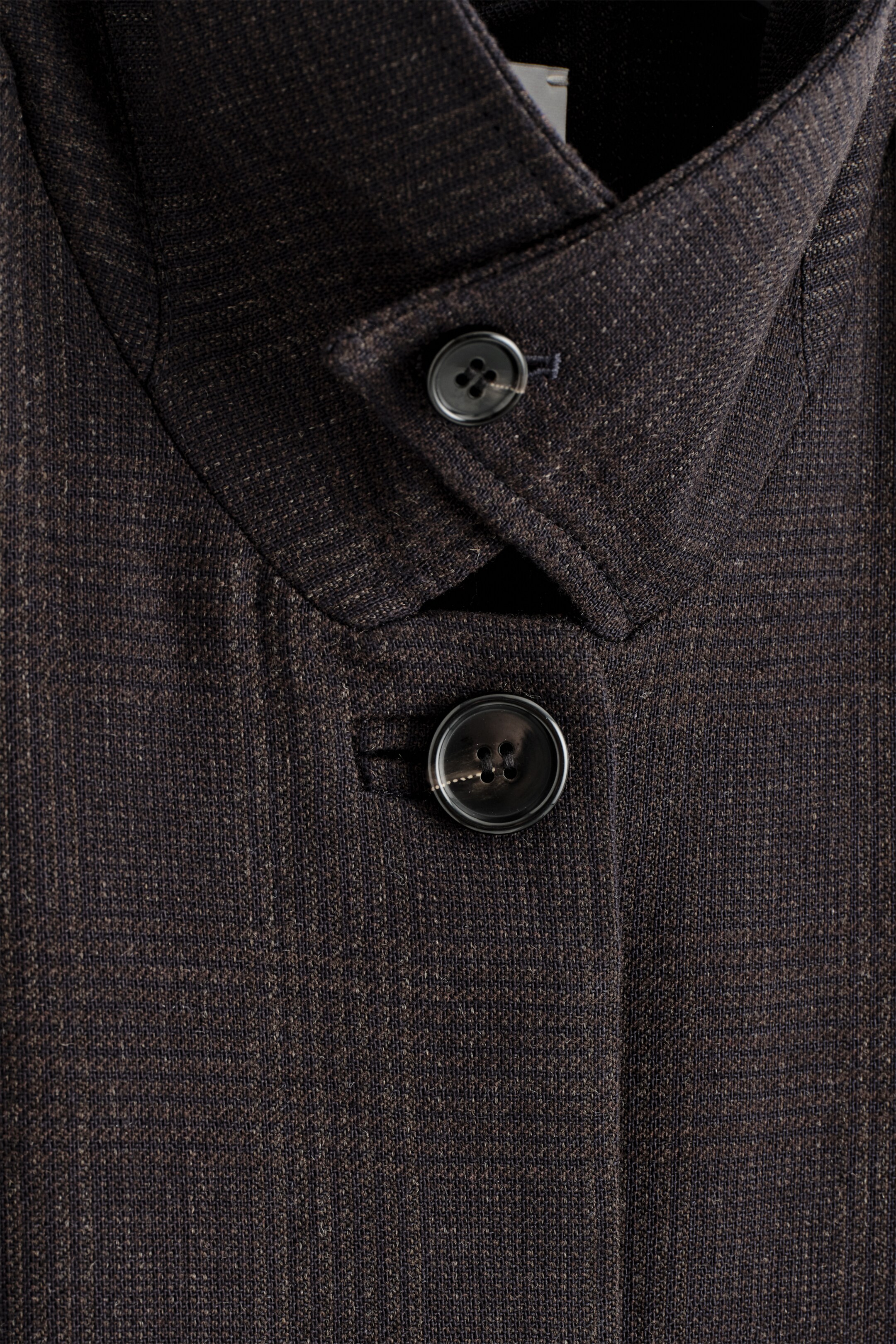 Agrandir l'image: Un manteau sombre, subtilement texturé, est présenté en gros plan sur sa partie supérieure avant, mettant en valeur son col haut et pointu ainsi que sa fermeture à boutons sur le devant. Deux boutons sombres ferment le manteau, avec une petite étiquette pendante du bouton inférieur.