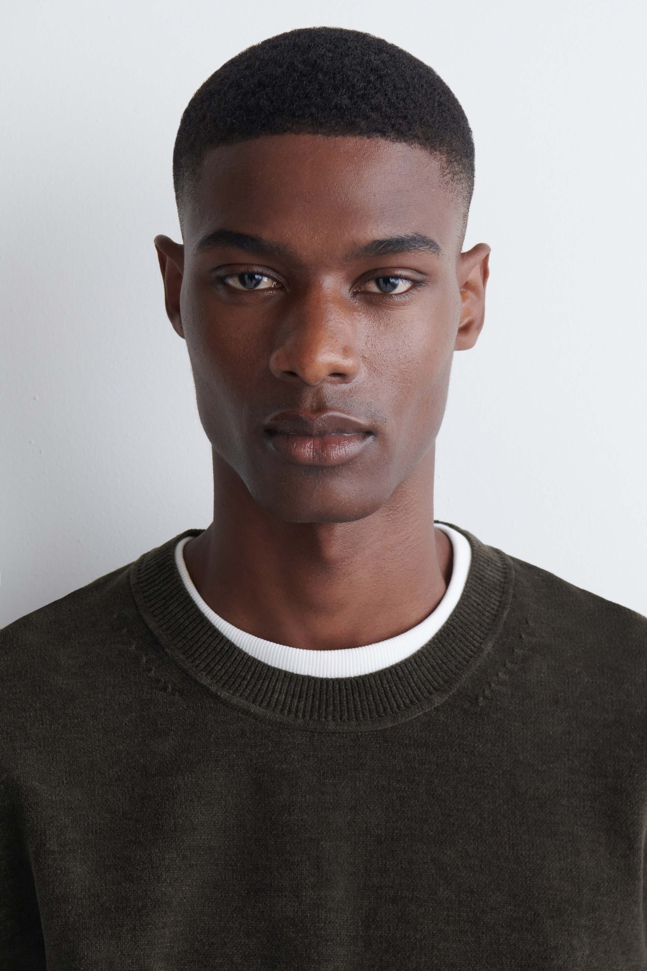 A dark green cotton-chenille crewneck sweater with a ribbed collar is being worn by the model. The sweater is styled with a white crewneck T-shirt layered underneath. A dark green cotton-chenille crewneck sweater with a ribbed collar is being worn by the model. The sweater is styled with a white crewneck T-shirt layered underneath.
