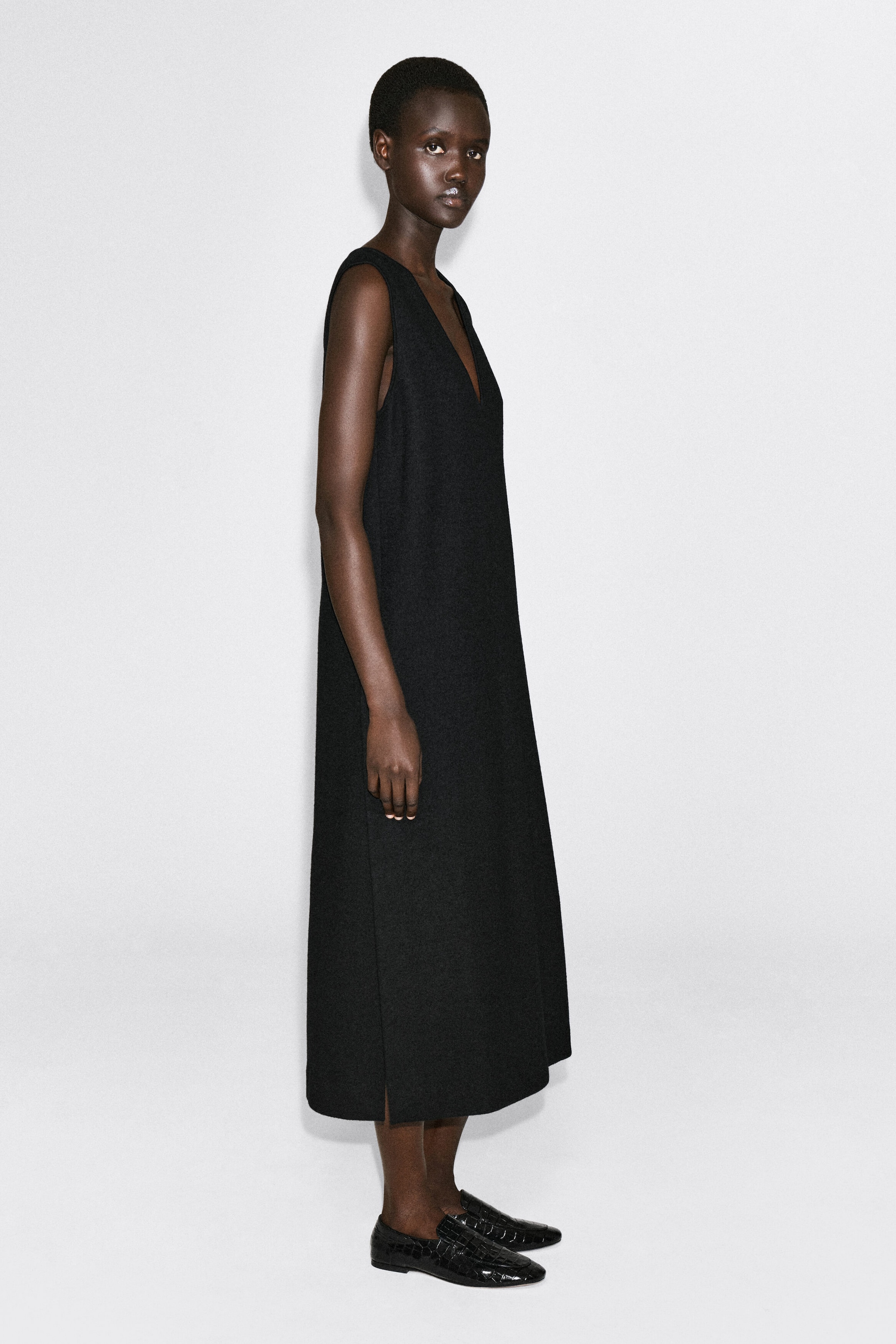 The black boiled wool-blend V-neck midi dress is shown on a model facing right, highlighting its sleeveless design and structured, straight-cut silhouette. The midi-length dress falls to mid-calf and features a deep V-neckline with a small side slit at the hem. The black boiled wool-blend V-neck midi dress is shown on a model facing right, highlighting its sleeveless design and structured, straight-cut silhouette. The midi-length dress falls to mid-calf and features a deep V-neckline with a small side slit at the hem.