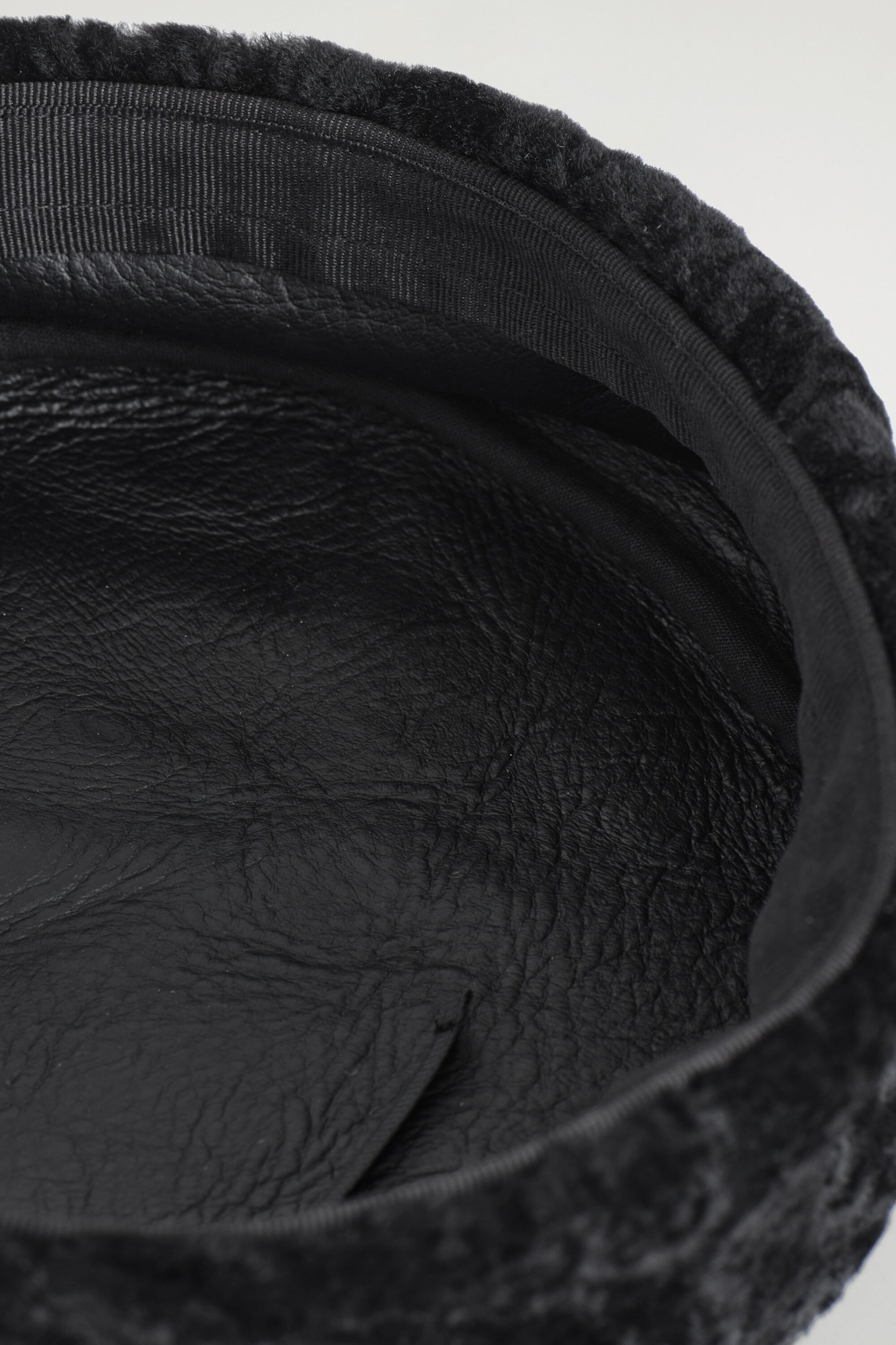 The underside interior of a black hat reveals a textured black leather lining and a ribbed fabric sweatband along the inner rim. Curly black shearling borders the outer edge of the hat. The underside interior of a black hat reveals a textured black leather lining and a ribbed fabric sweatband along the inner rim. Curly black shearling borders the outer edge of the hat.