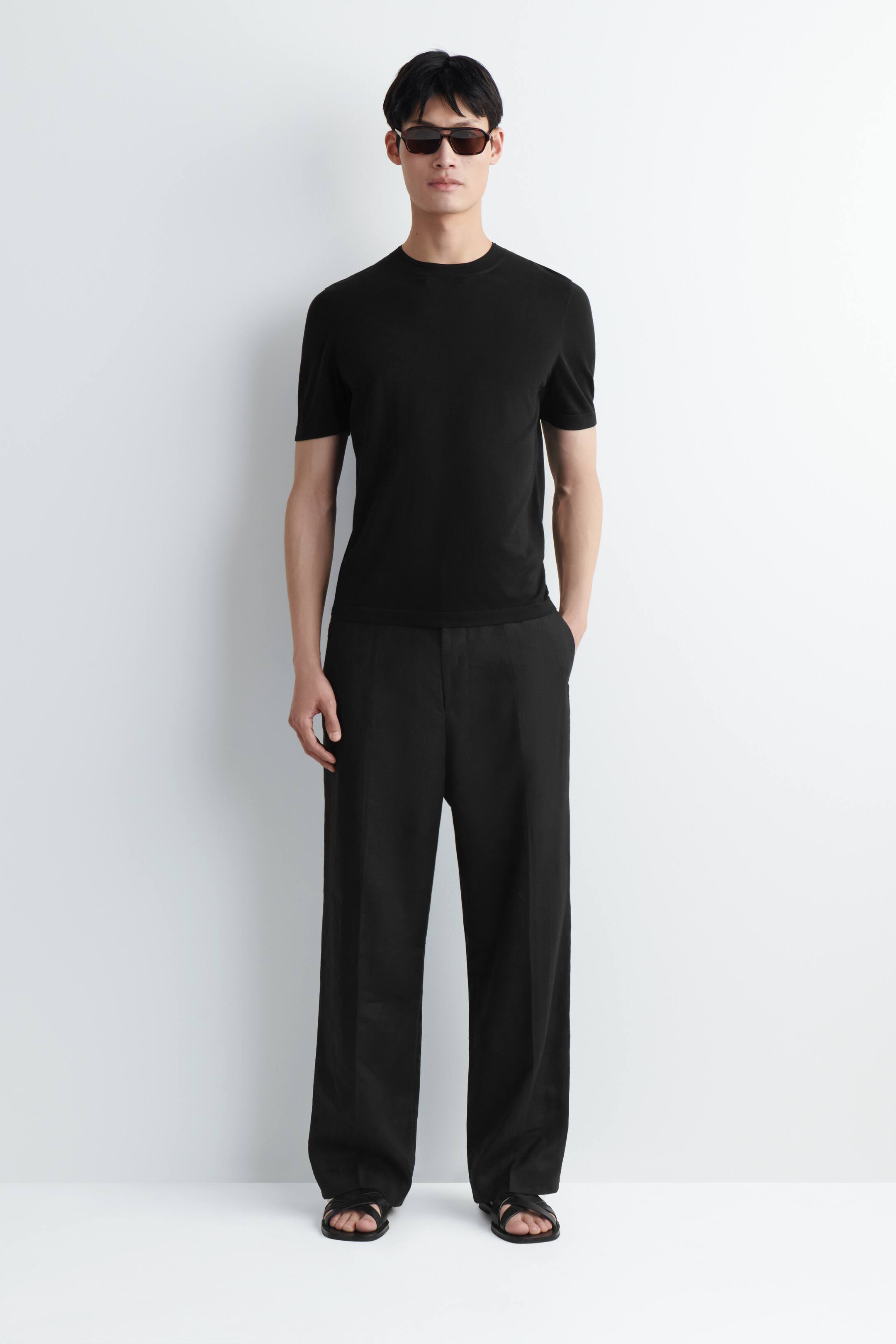 Vezi imaginea mai mare: Un tricou negru cu mânecă scurtă și guler rotund este purtat cu pantaloni negri, largi, cu croială lejeră și lungime completă. Ținuta este completată cu ochelari de soare cu rame închise la culoare și sandale negre cu barete plate.