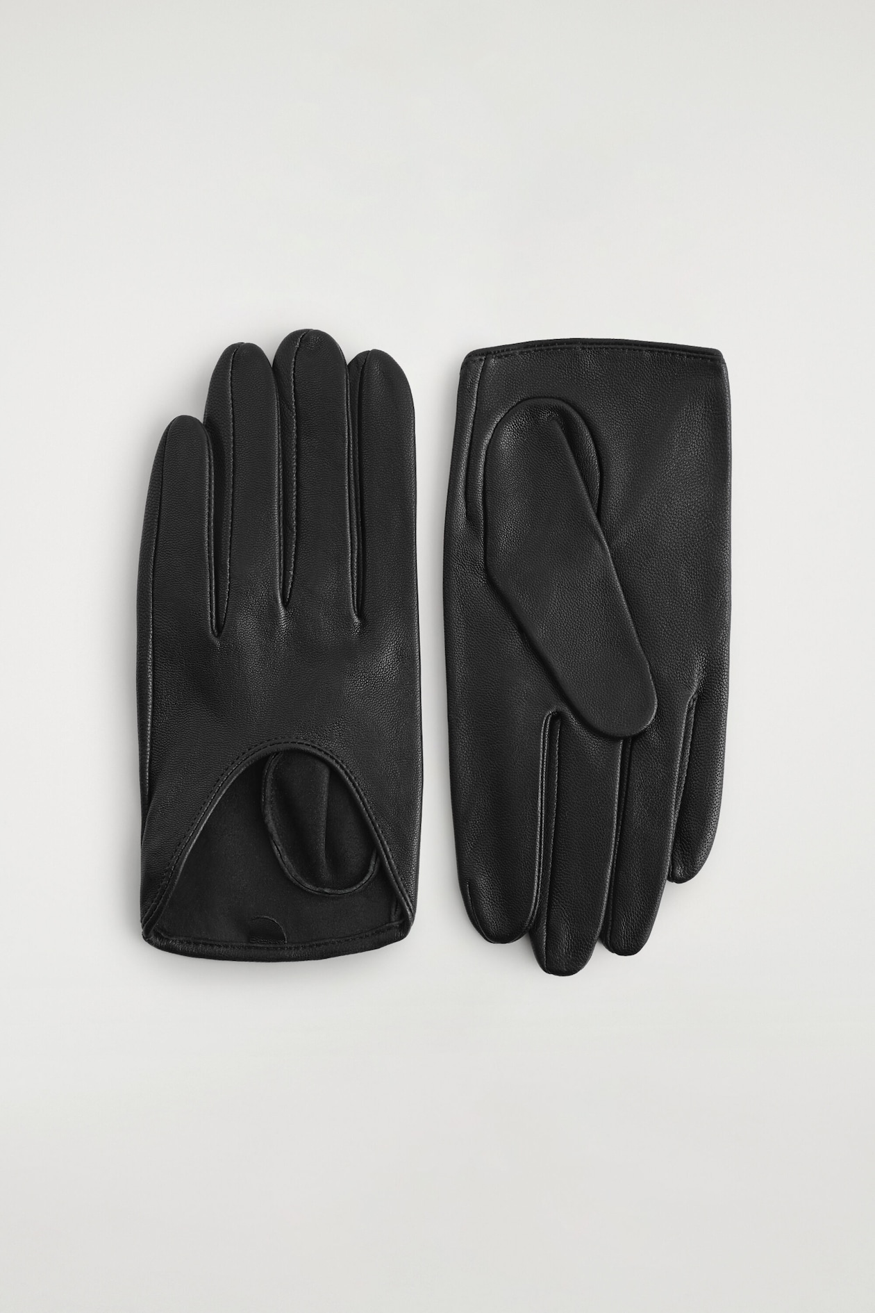 A pair of black leather driving gloves lies flat, displaying a textured grain finish with tonal stitching. The glove on the left shows the back of the hand with a classic keyhole cutout at the wrist, while the glove on the right shows the palm side with a folded thumb.