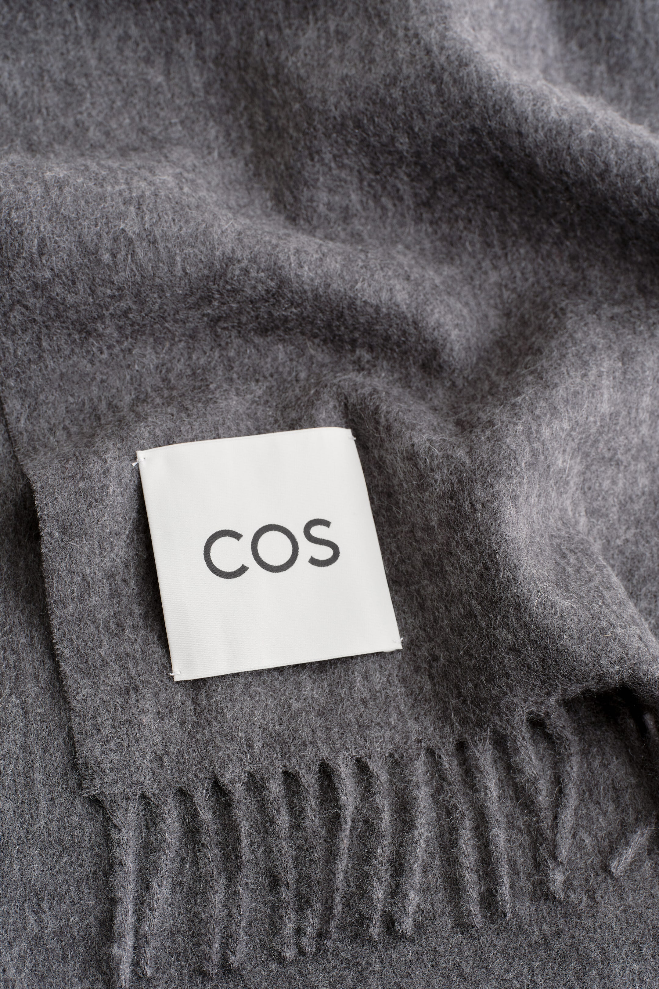 A dark grey fringed cashmere scarf is shown in close-up, highlighting its soft, brushed texture and twisted fringe detailing along the edge. A square white fabric label featuring the black “COS” brand name is stitched onto the front corner of the draped fabric. A dark grey fringed cashmere scarf is shown in close-up, highlighting its soft, brushed texture and twisted fringe detailing along the edge. A square white fabric label featuring the black “COS” brand name is stitched onto the front corner of the draped fabric.