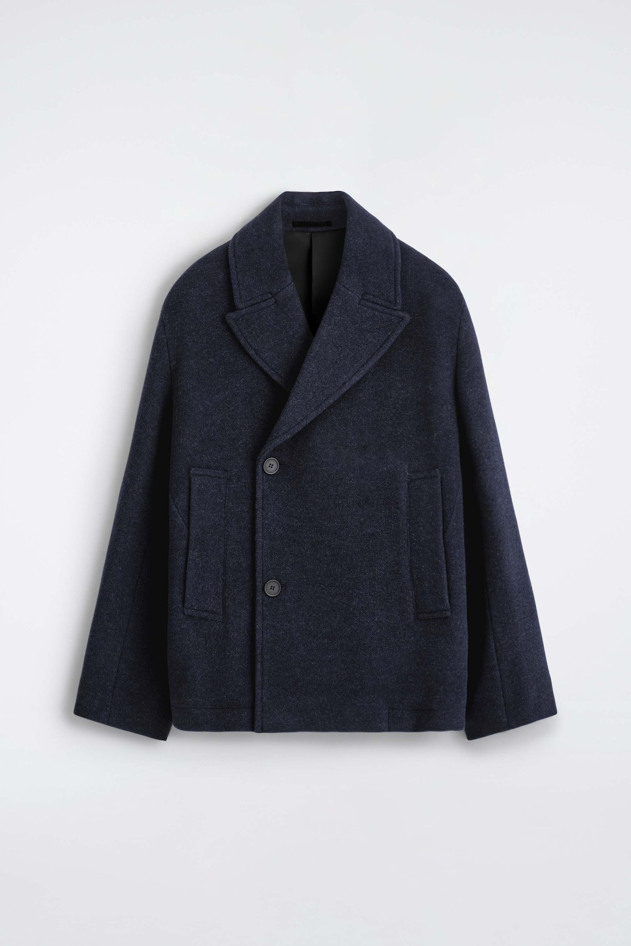A double-breasted wool short pea coat in dark navy is shown from the front, showcasing its wide notch lapels and structured, boxy silhouette. It features a double-breasted fastening with two dark buttons, long relaxed sleeves, and vertical welt pockets on each side.