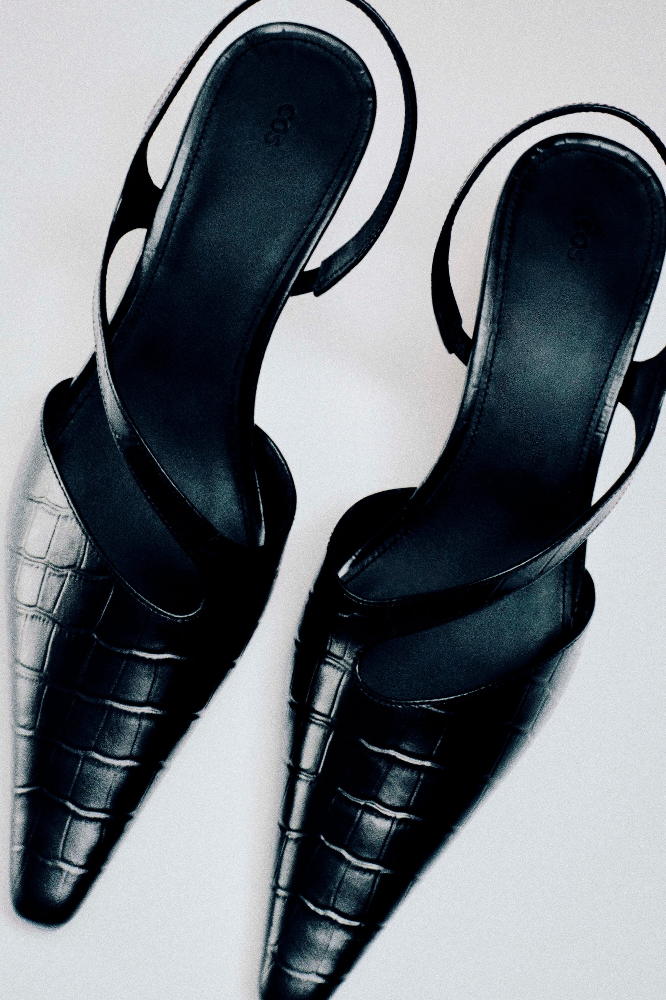 These black slingback pumps showcase a pointed-toe silhouette crafted from croc-embossed leather. They feature asymmetrical cut-out straps that curve over the upper foot and thin slingback straps, viewed from a top-down angle.