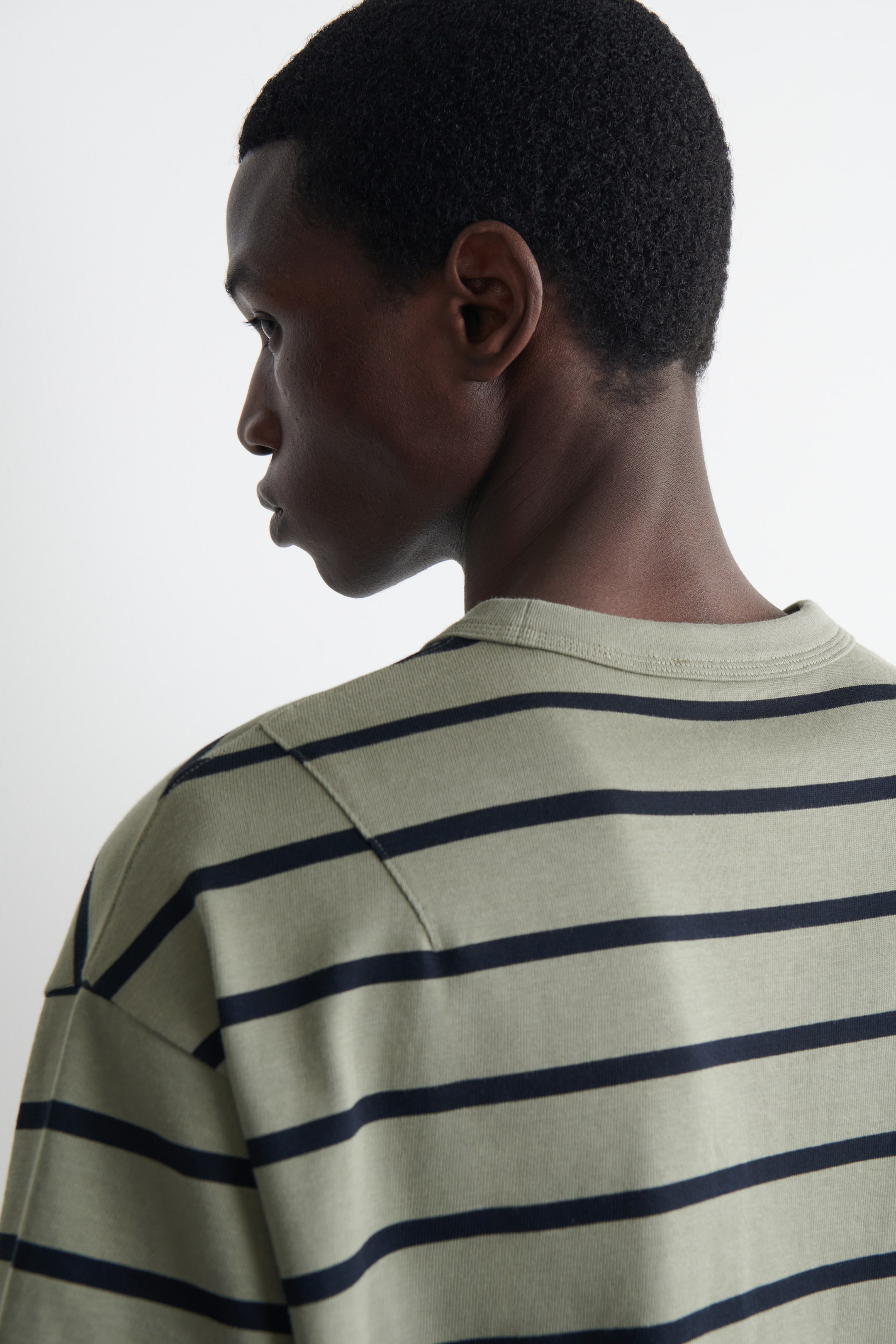 Agrandir l'image: Un t-shirt vert olive clair avec des rayures horizontales bleu marine est porté, vu de dos et du côté gauche. Il présente un col rond et une couture d'épaule visible à travers le haut du dos.