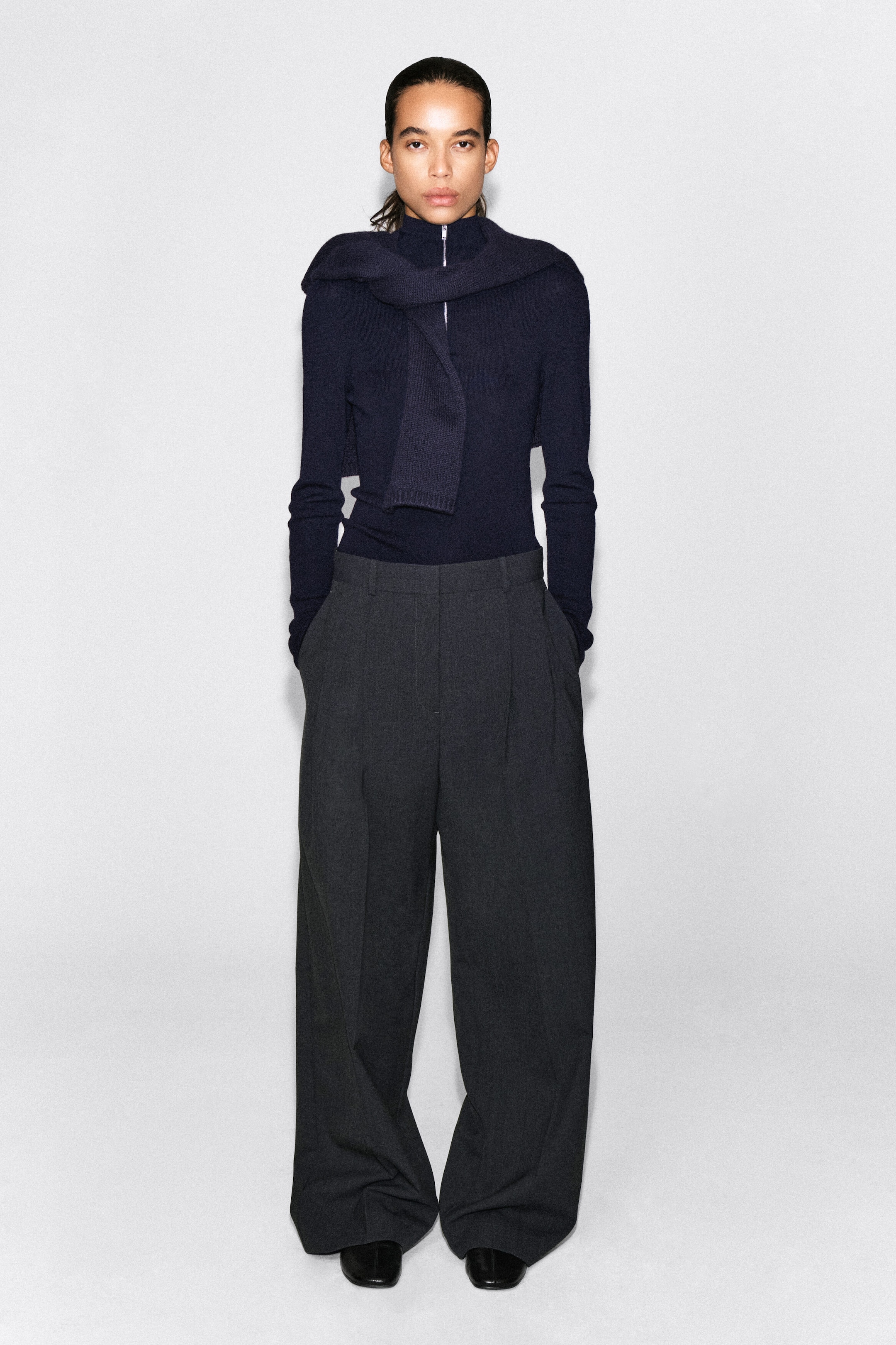 A navy blue half-zip knit jumper is being worn with a matching draped scarf and dark grey wide-leg trousers, shown from the front. The high-waisted trousers fall loosely with front pleats, while the slim-fit jumper is tucked in to highlight the structured waistline. A navy blue half-zip knit jumper is being worn with a matching draped scarf and dark grey wide-leg trousers, shown from the front. The high-waisted trousers fall loosely with front pleats, while the slim-fit jumper is tucked in to highlight the structured waistline.