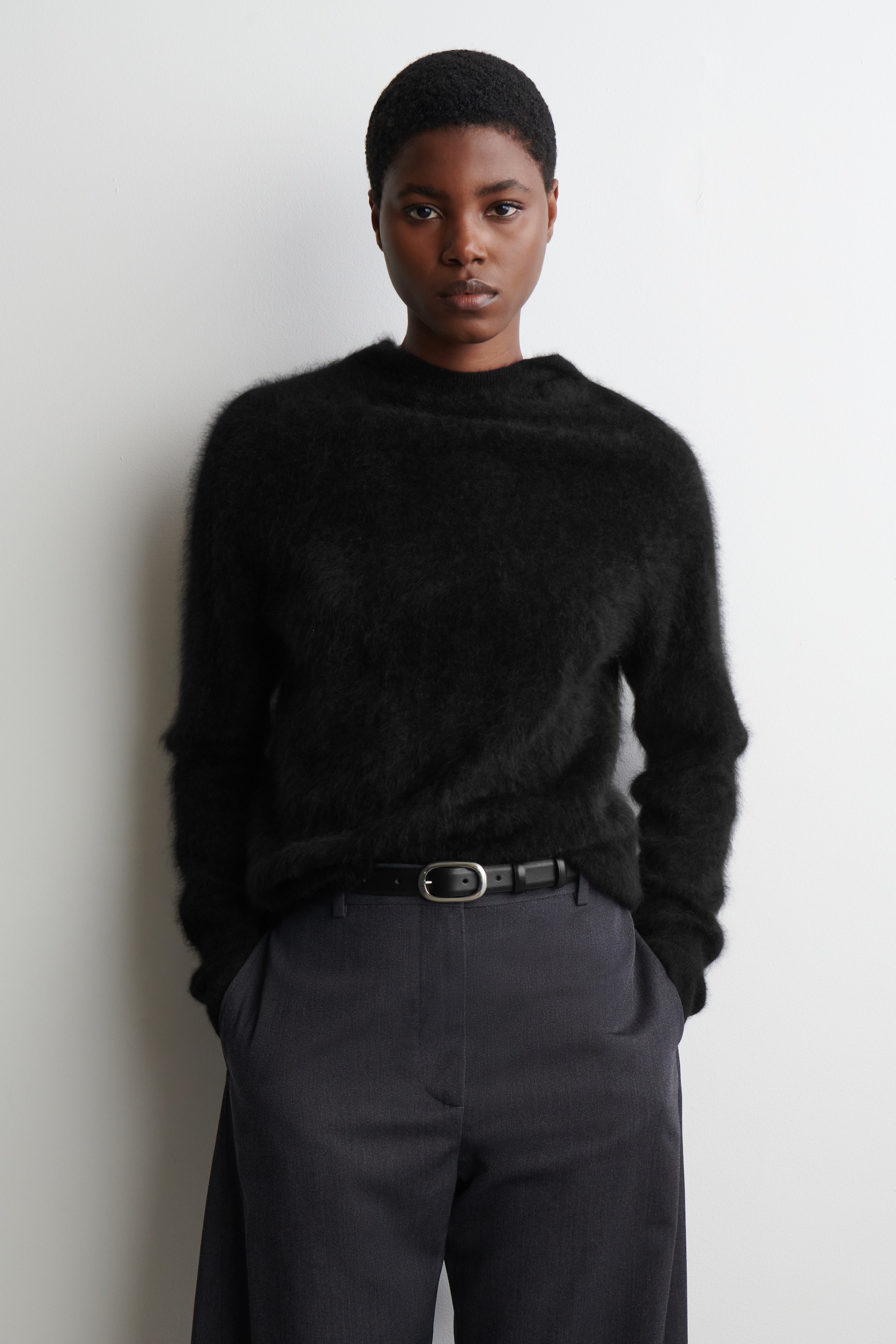View larger image: A black fluffy sweater with a high crew neck and long sleeves is being presented, showcasing its relaxed fit and subtle front tuck into grey trousers.