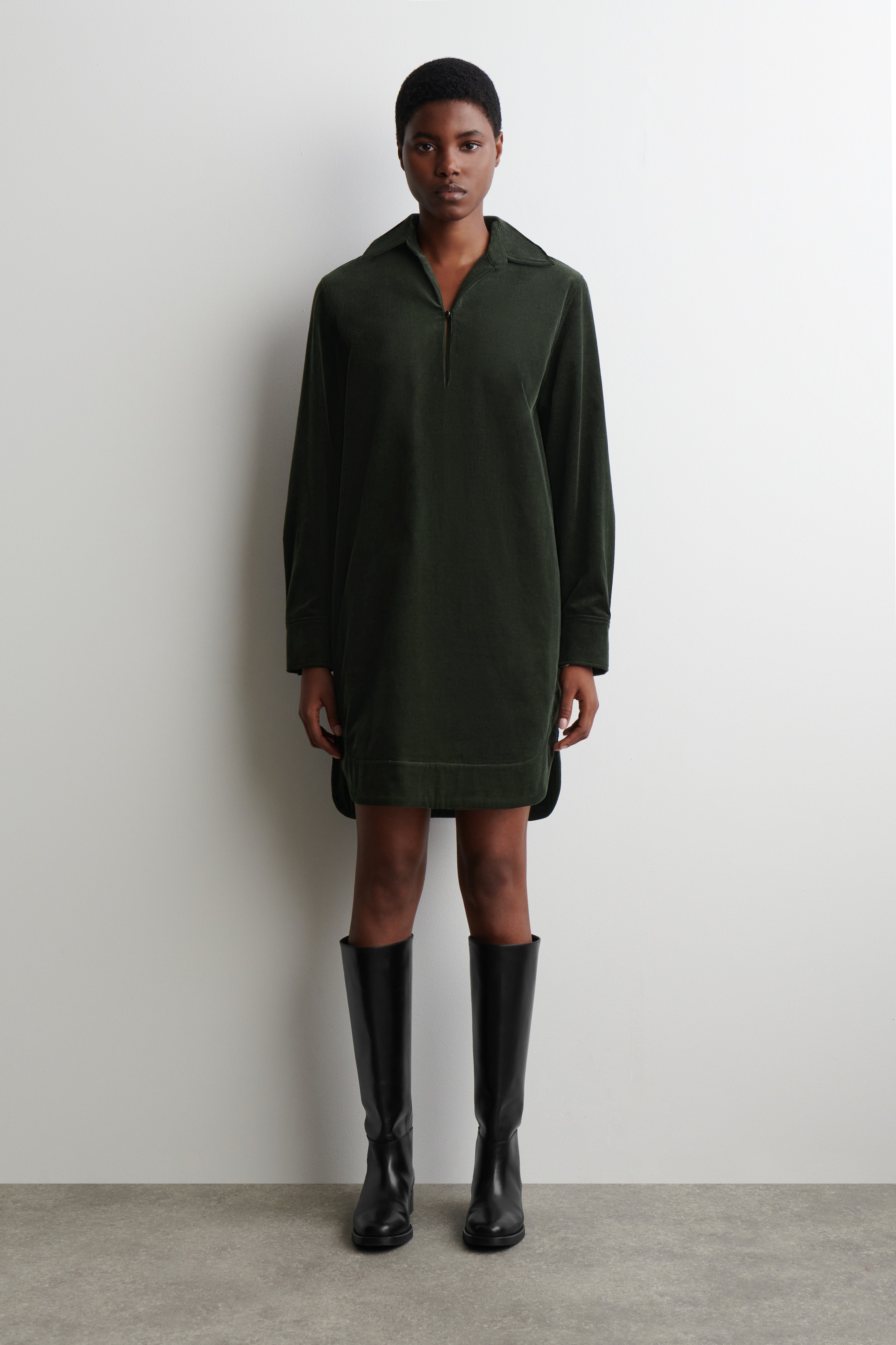 Vezi imaginea mai mare: O rochie de culoare verde închis, asemănătoare cu velurul, este orientată frontal, având un guler polo cu o deschidere parțială cu fermoar în față. Prezentând mâneci lungi și o croială dreaptă, relaxată, aceasta se termină cu un tiv larg deasupra genunchiului.