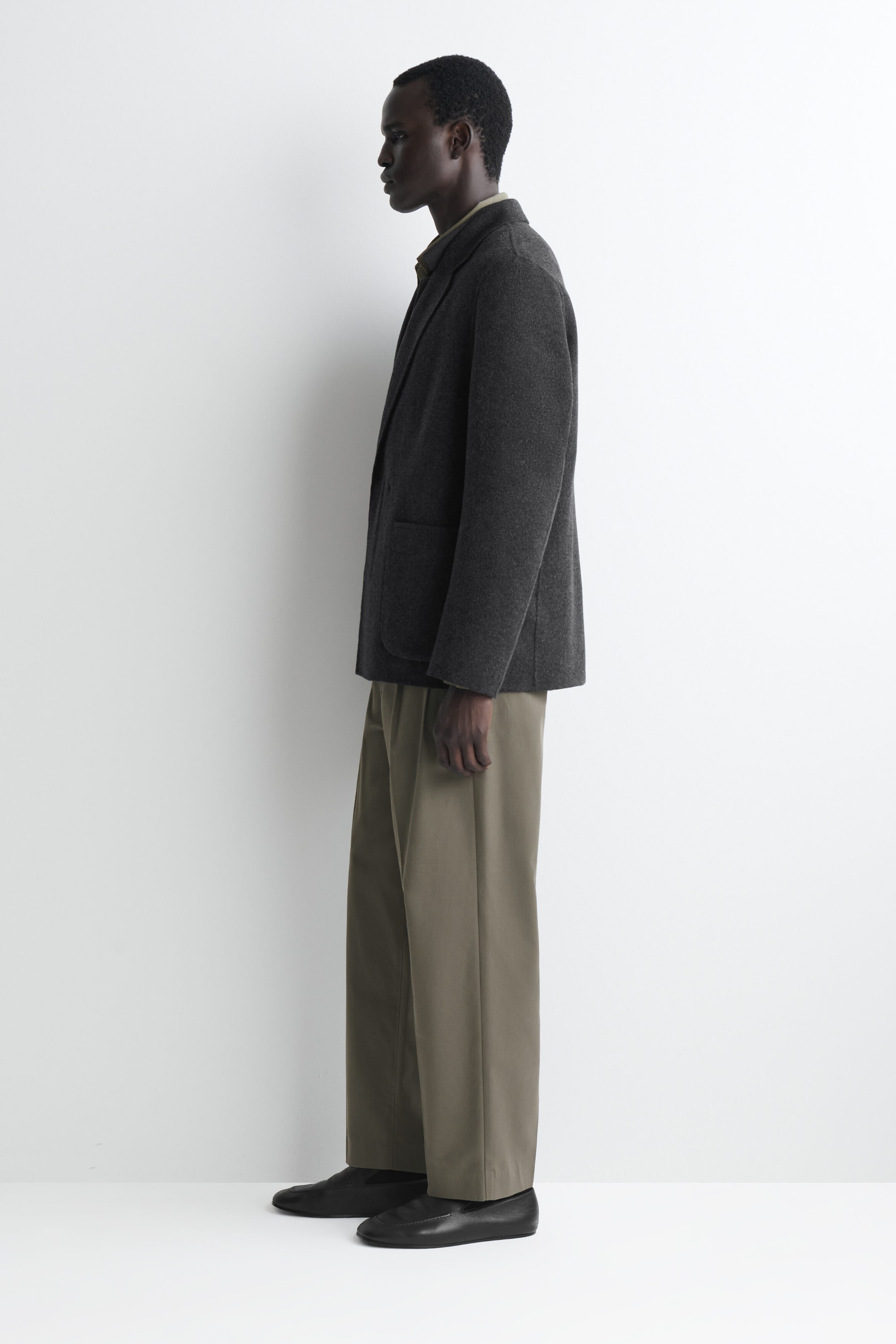 A dark grey unstructured wool-blend blazer is worn in side profile facing left, showcasing its relaxed fit and soft drape. The blazer features a patch pocket and is paired with wide-leg olive trousers and black leather loafers. A dark grey unstructured wool-blend blazer is worn in side profile facing left, showcasing its relaxed fit and soft drape. The blazer features a patch pocket and is paired with wide-leg olive trousers and black leather loafers.