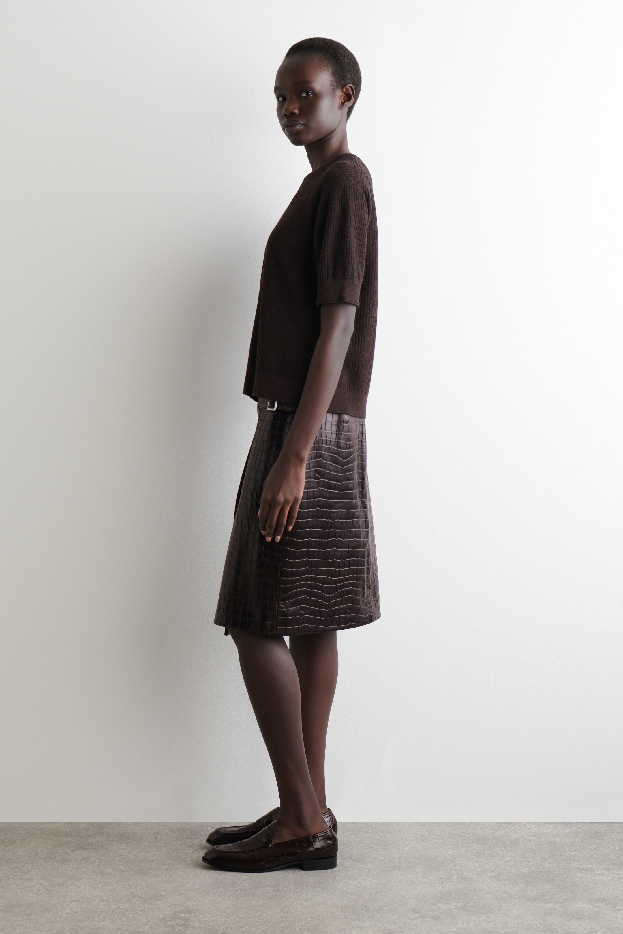 View larger image: A dark brown ribbed knit top with short sleeves and a round neckline is being worn with a dark brown knee-length skirt, featuring an embossed crocodile pattern and cinched with a thin belt. Dark brown loafers with a textured finish are completing the outfit, all viewed from a side profile.
