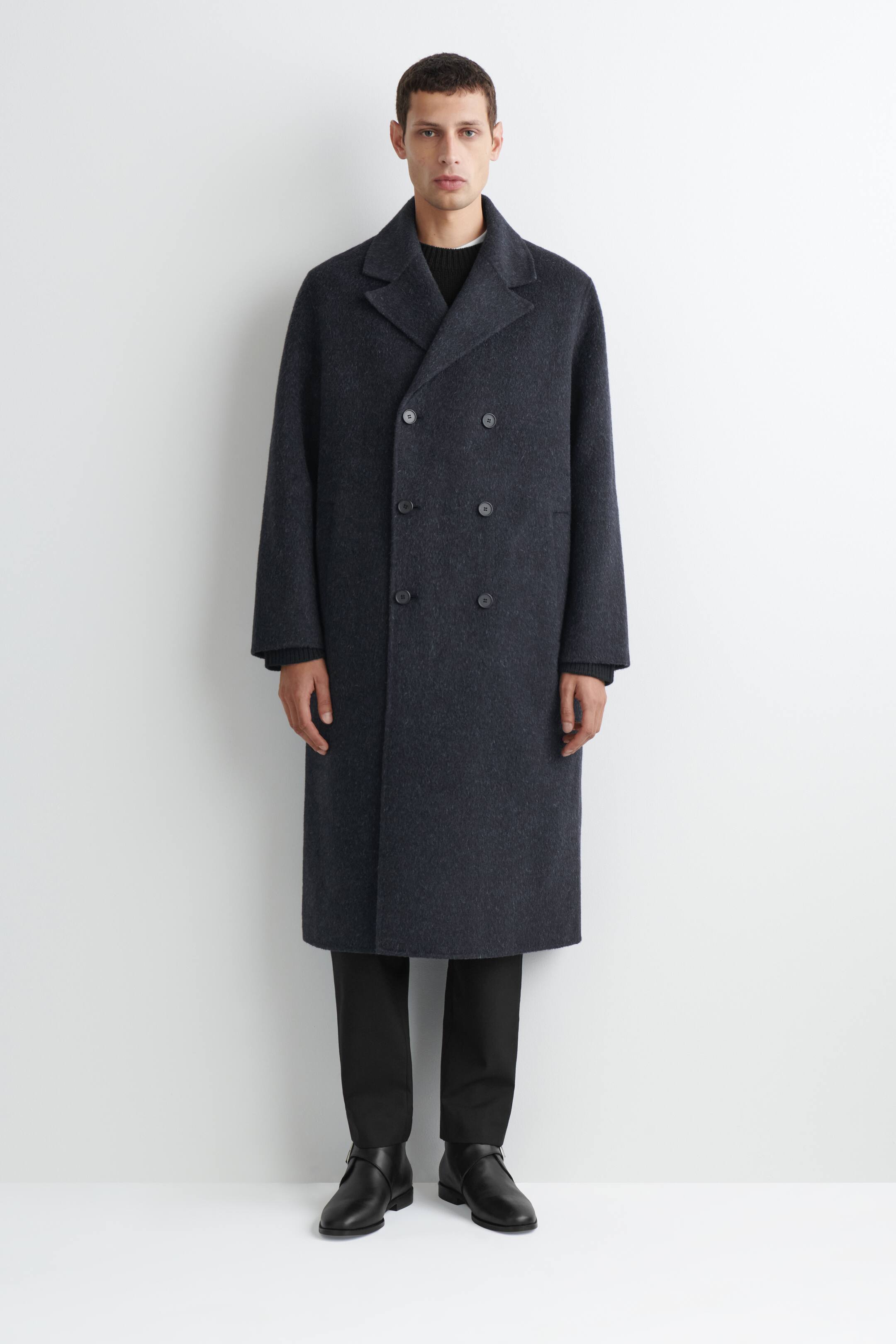 View larger image: The double-faced wool-blend long coat is shown from the front in a relaxed, straight-cut silhouette, featuring a double-breasted button closure and classic notch lapels in dark charcoal grey. The knee-length coat is layered over a dark crew-neck sweater and black trousers to showcase its drape and structure.