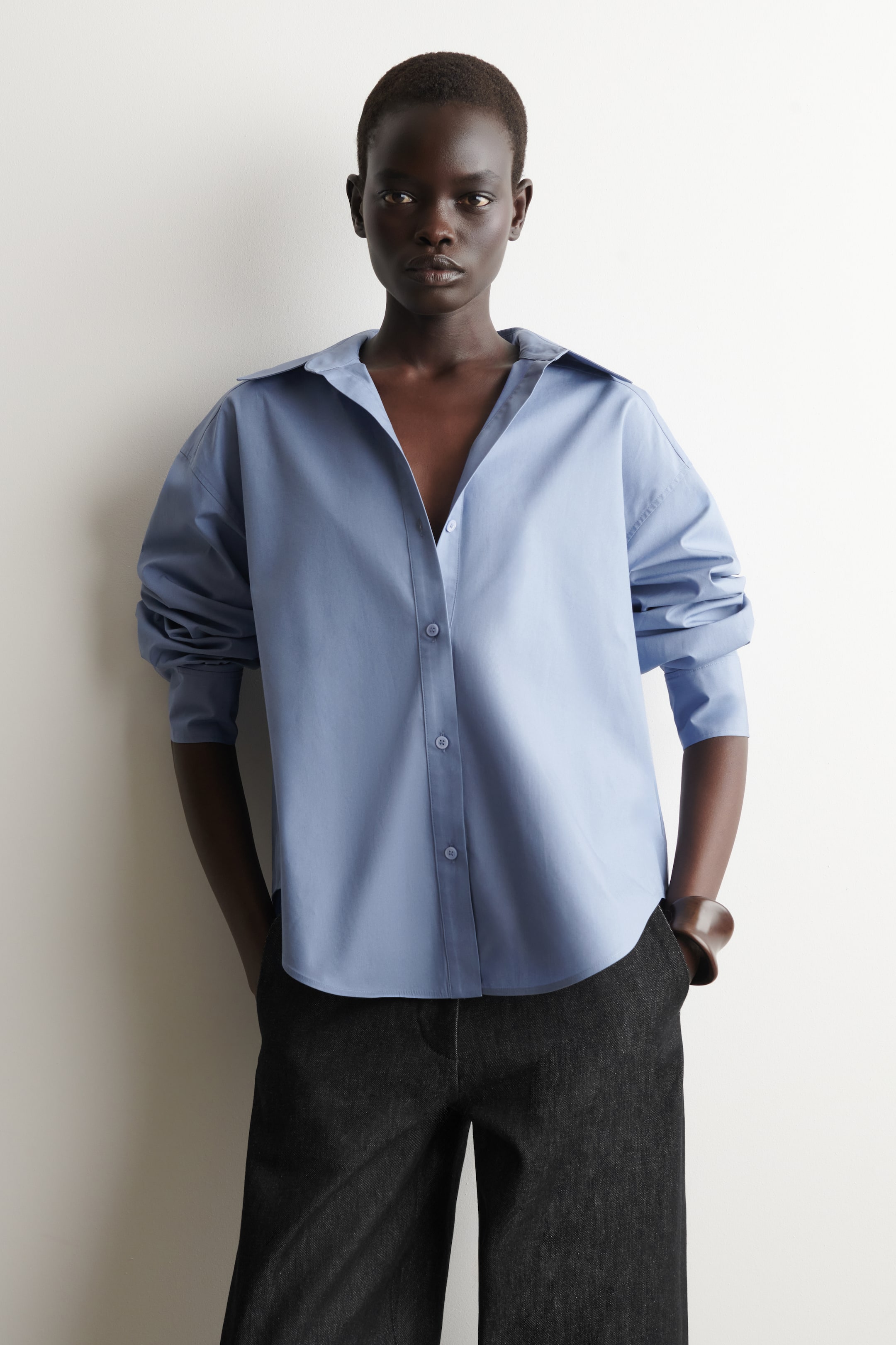 Zobacz większy obraz: Jasnoniebieska koszula zapinana na guziki jest prezentowana z luźnym krojem, opadającymi ramionami i rozłożystym kołnierzem noszonym otwartym. Długie rękawy są podwinięte, a zakrzywiony dół koszuli pozostaje nie włożony w ciemne spodnie.