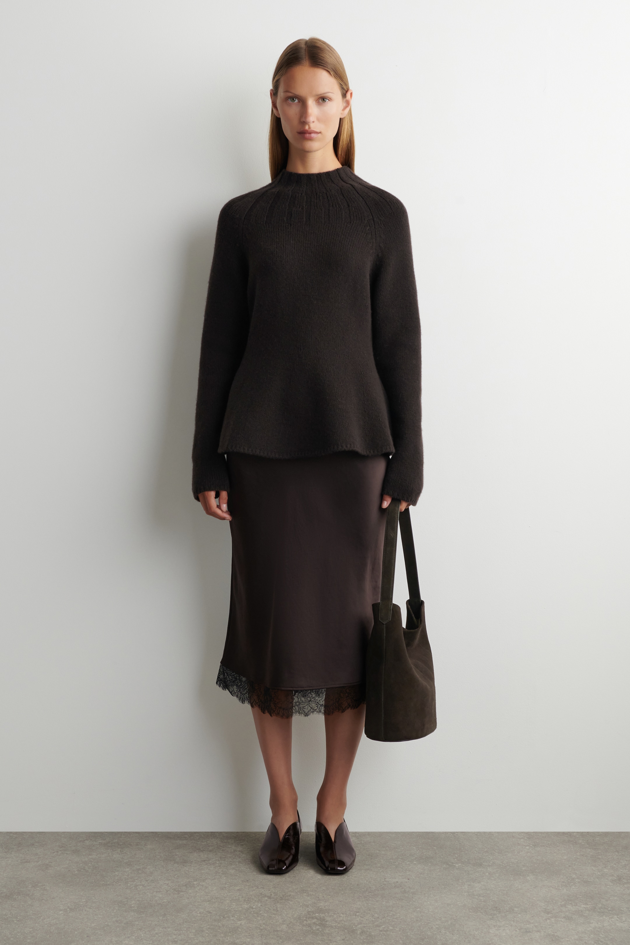 Agrandir l'image: Un pull col montant marron foncé, avec des coutures raglan et un ourlet légèrement évasé, est porté par-dessus une jupe midi en satin marron foncé avec une garniture en dentelle noire visible à l'ourlet. Un sac seau en daim marron foncé est tenu sur le côté, et des ballerines en cuir verni foncé avec un décolleté en forme de V complètent l'ensemble.