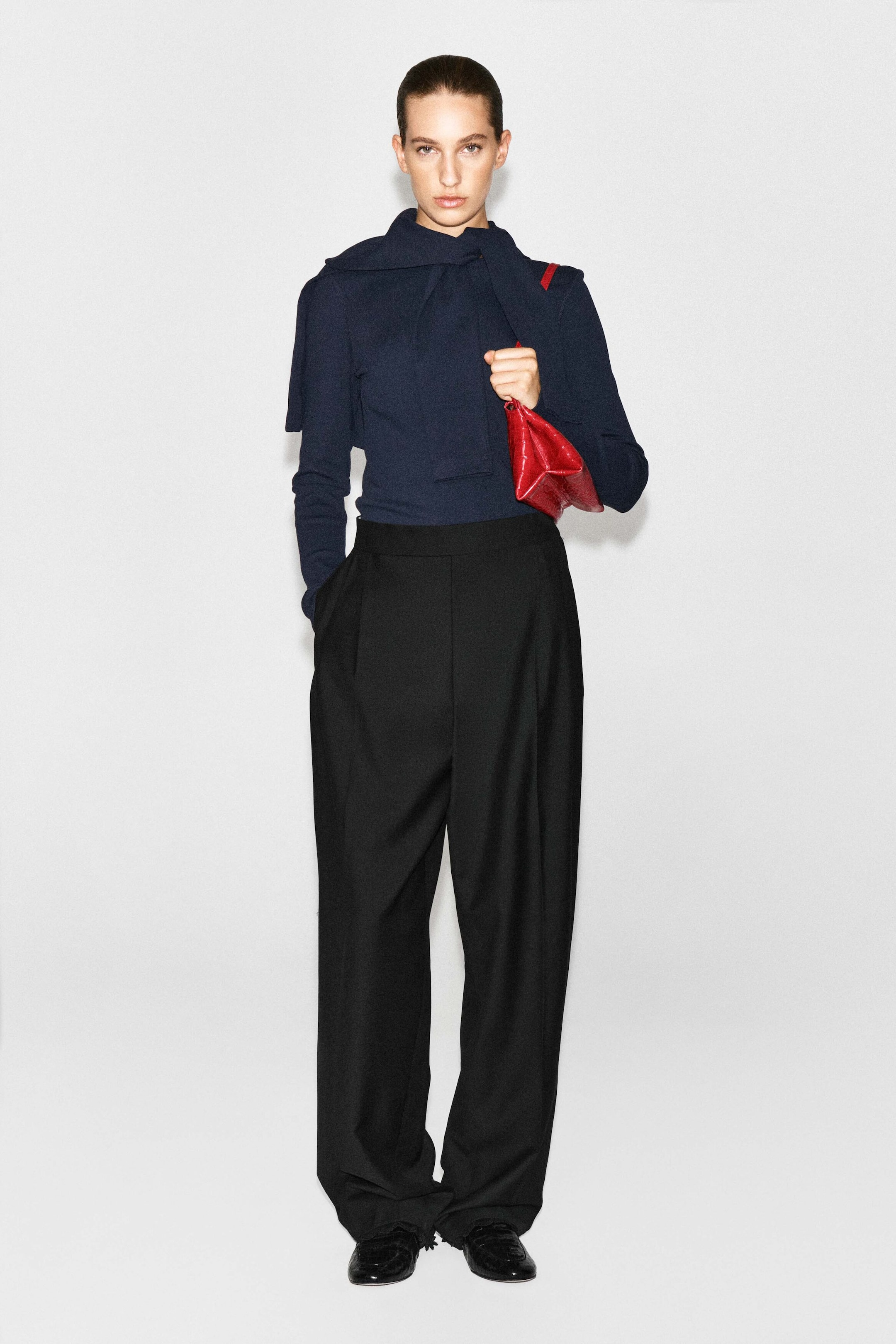 A navy blue slim ribbed cotton long-sleeved top with a draped neck-tie detail is being worn by the model. The top is tucked into wide-leg black trousers, with a small red handbag being held. A navy blue slim ribbed cotton long-sleeved top with a draped neck-tie detail is being worn by the model. The top is tucked into wide-leg black trousers, with a small red handbag being held.
