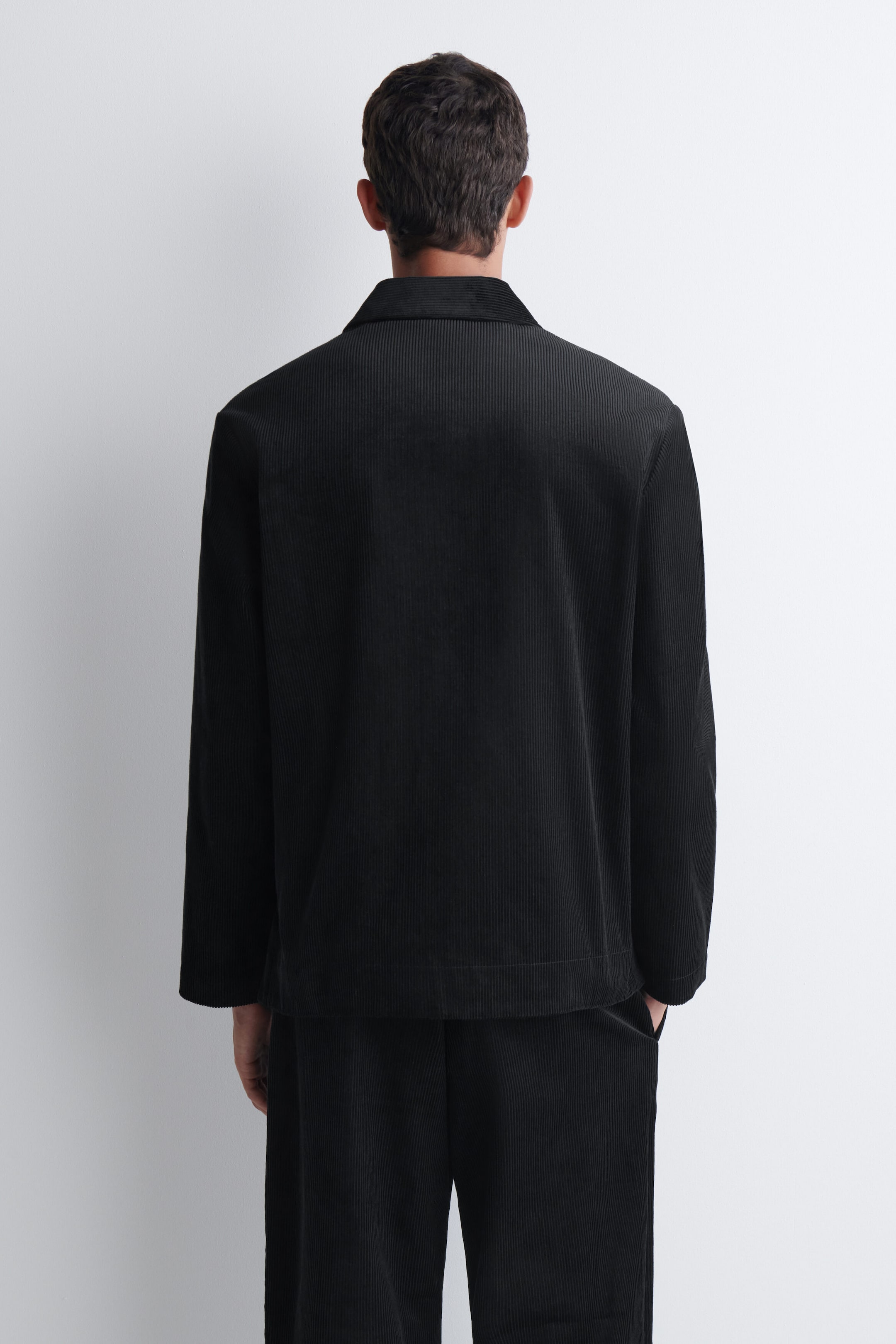 Une veste en velours côtelé noir avec col et manches longues est portée par le mannequin, vue de dos. La veste est assortie à un pantalon en velours côtelé noir.