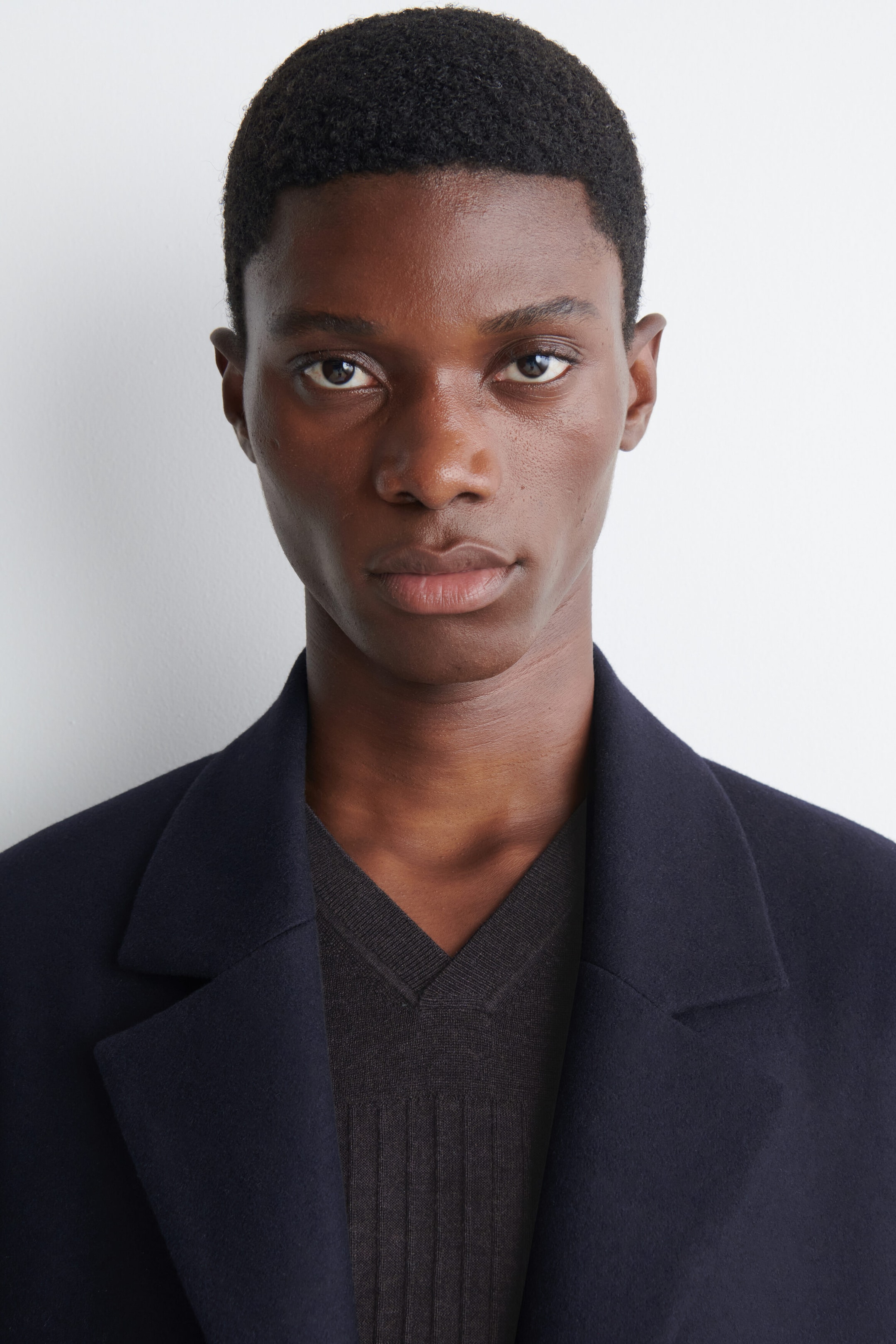 The model is wearing a dark navy tailored coat with wide lapels. It is layered over a dark grey V-neck jumper with a ribbed texture. The model is wearing a dark navy tailored coat with wide lapels. It is layered over a dark grey V-neck jumper with a ribbed texture.