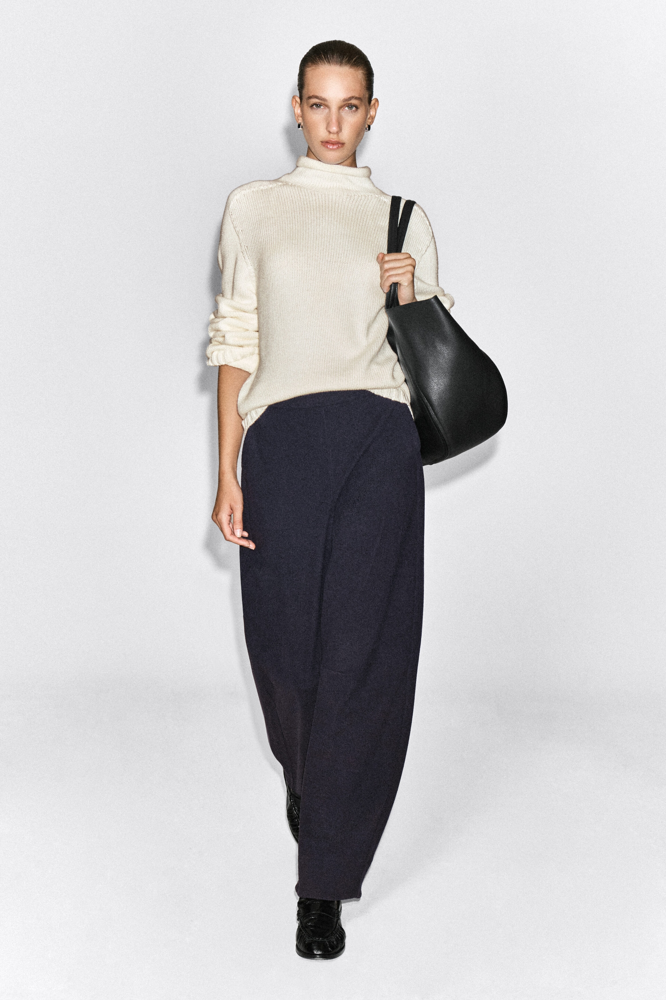 The model is wearing dark navy blue corduroy-jersey wide-leg pants, showcasing a relaxed silhouette and soft texture. The pants are paired with a cream-colored high-neck knit sweater and black loafers.