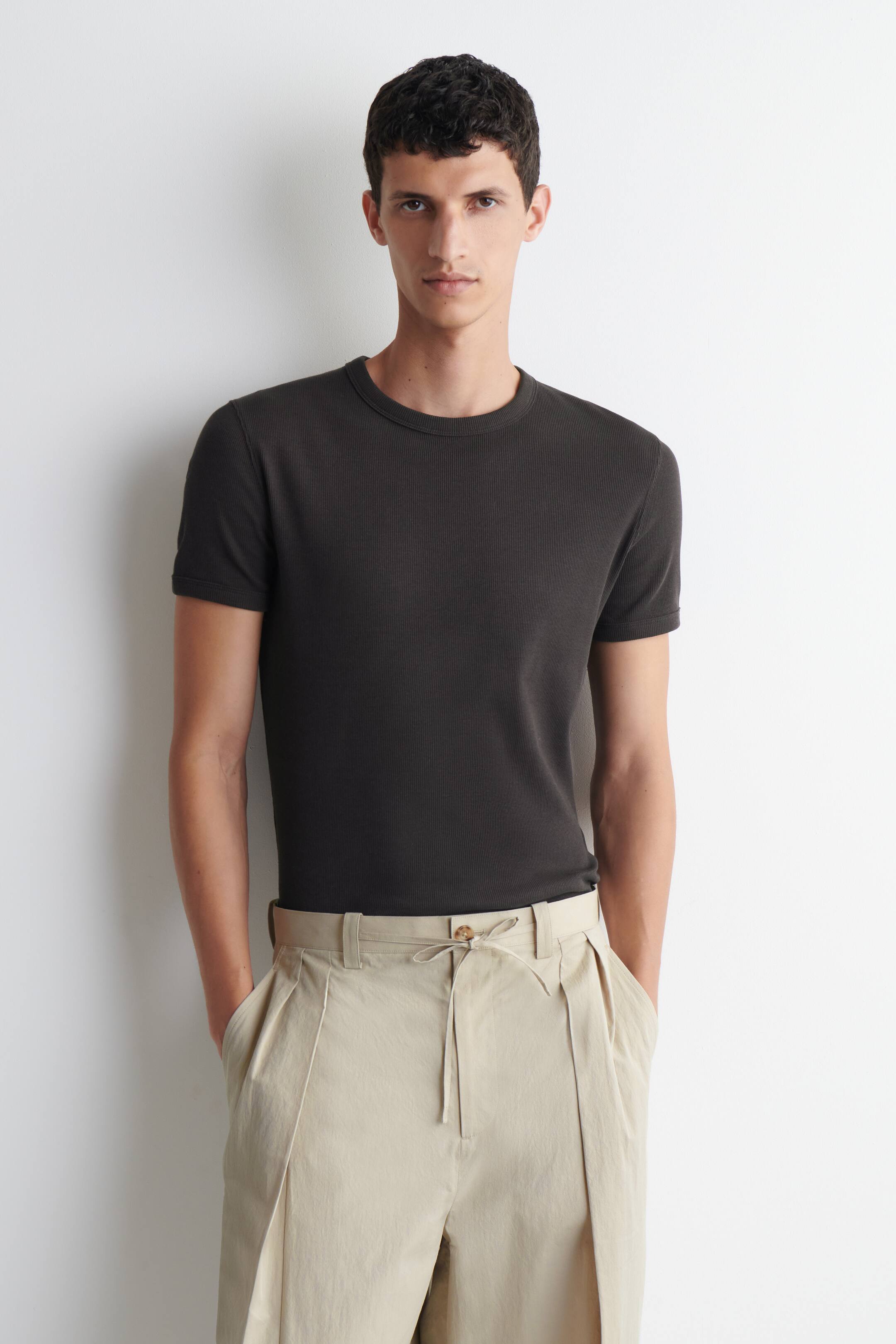 Größeres Bild ansehen: Ein dunkelgraues, geripptes T-Shirt mit Rundhalsausschnitt und kurzen Ärmeln wird vom Model getragen und liegt schlank am Oberkörper und an den Armen an. Das T-Shirt ist mit hellen, plissierten Hosen kombiniert.