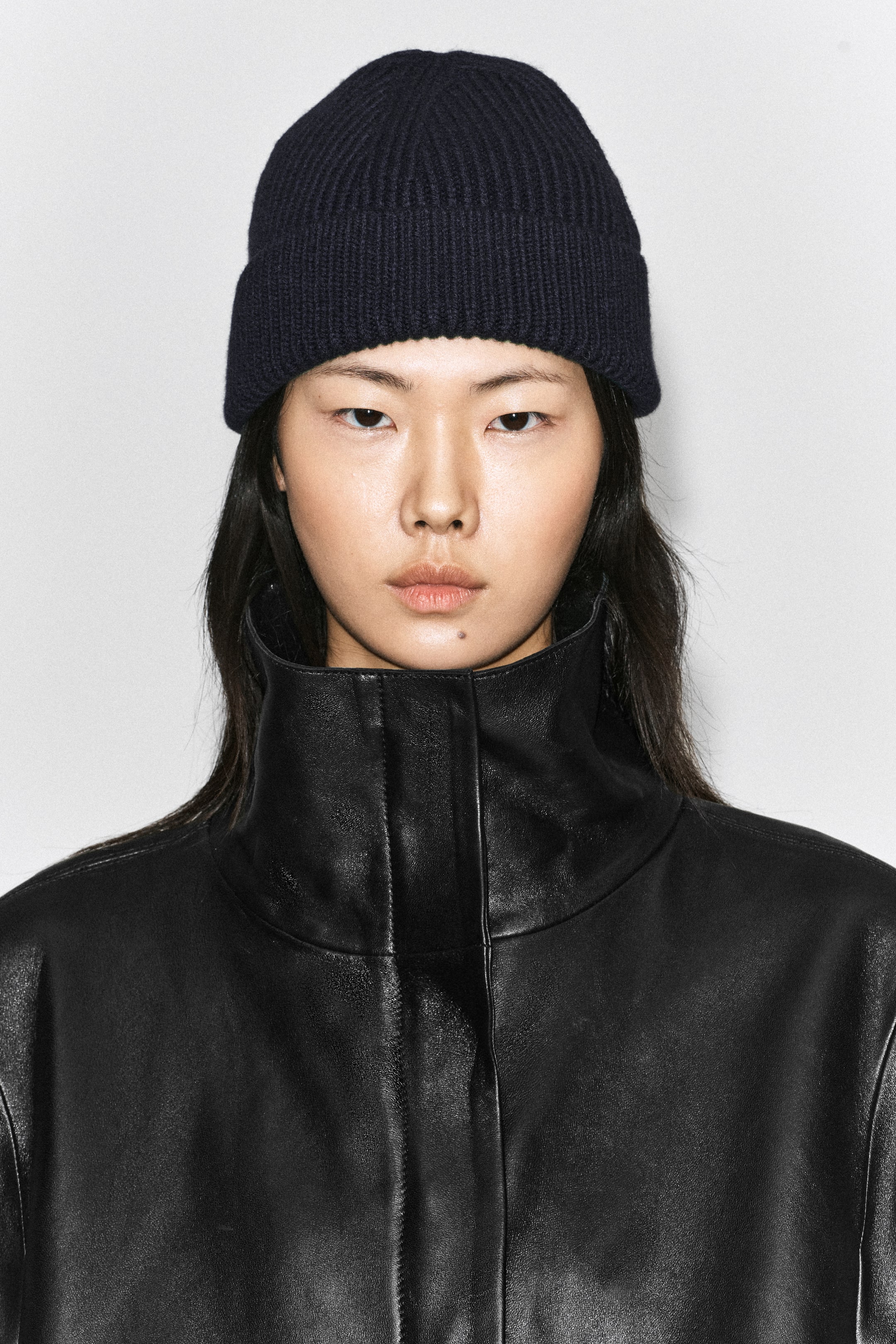 Agrandir l'image: Un bonnet en laine côtelée épaisse bleu marine foncé est porté par le mannequin, avec un large revers replié. Le bonnet est associé à une veste en cuir noire structurée à col montant.