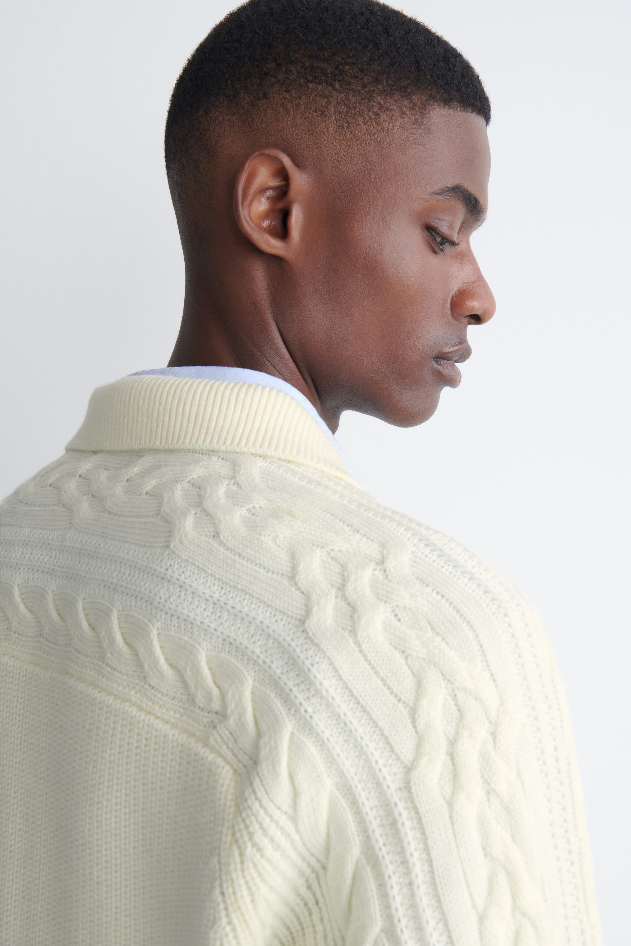 A cream-colored, chunky cable-knit polo sweater is being worn by the model, showcasing textured knit patterns across the shoulders and upper back. The sweater features a ribbed collar and is layered over a light blue shirt. A cream-colored, chunky cable-knit polo sweater is being worn by the model, showcasing textured knit patterns across the shoulders and upper back. The sweater features a ribbed collar and is layered over a light blue shirt.