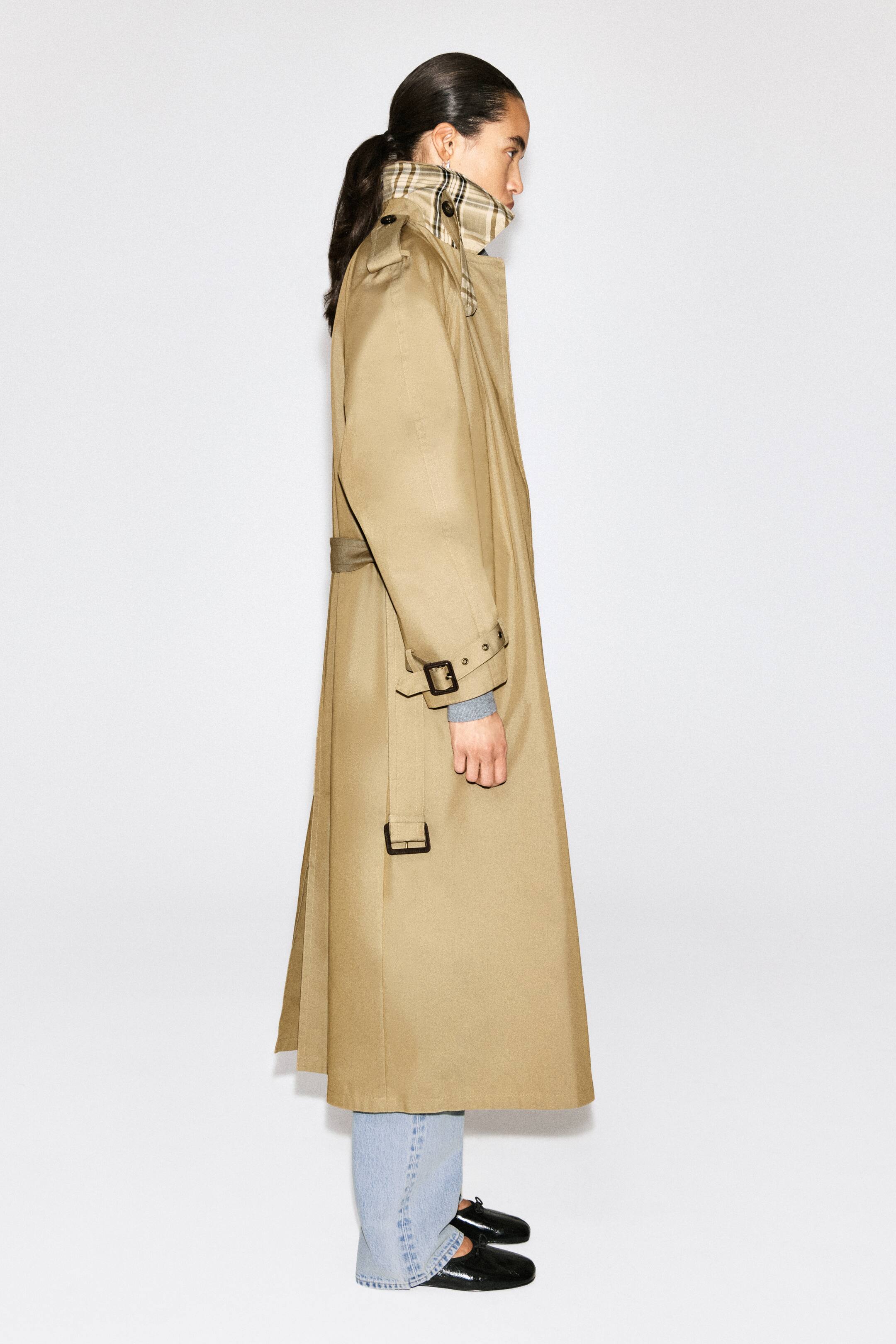 Agrandir l'image: Le trench-coat en coton ceinturé CHECK-DETAIL est présenté de profil gauche, en tissu coton beige, avec un col relevé laissant apparaître un motif à carreaux, des pattes d’épaule et des brides de poignet à boucle. Une ceinture assortie souligne la taille, et le manteau, de longueur longue, recouvre partiellement un pull gris au poignet ainsi qu’un jean délavé clair à l’ourlet.
