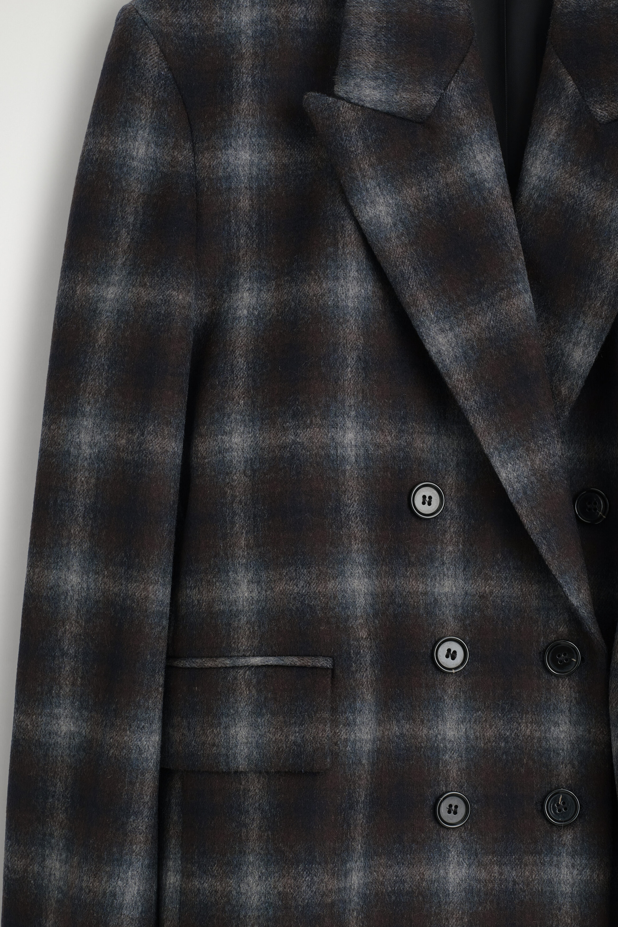 Un blazer croisé en mélange de laine à carreaux est présenté de face dans un gros plan, mettant en valeur son motif écossais gris foncé et marron ainsi que sa texture douce en mélange de laine. Le blazer affiche de larges revers en pointe, une fermeture croisée à double rangée de boutons et une poche à rabat structurée.