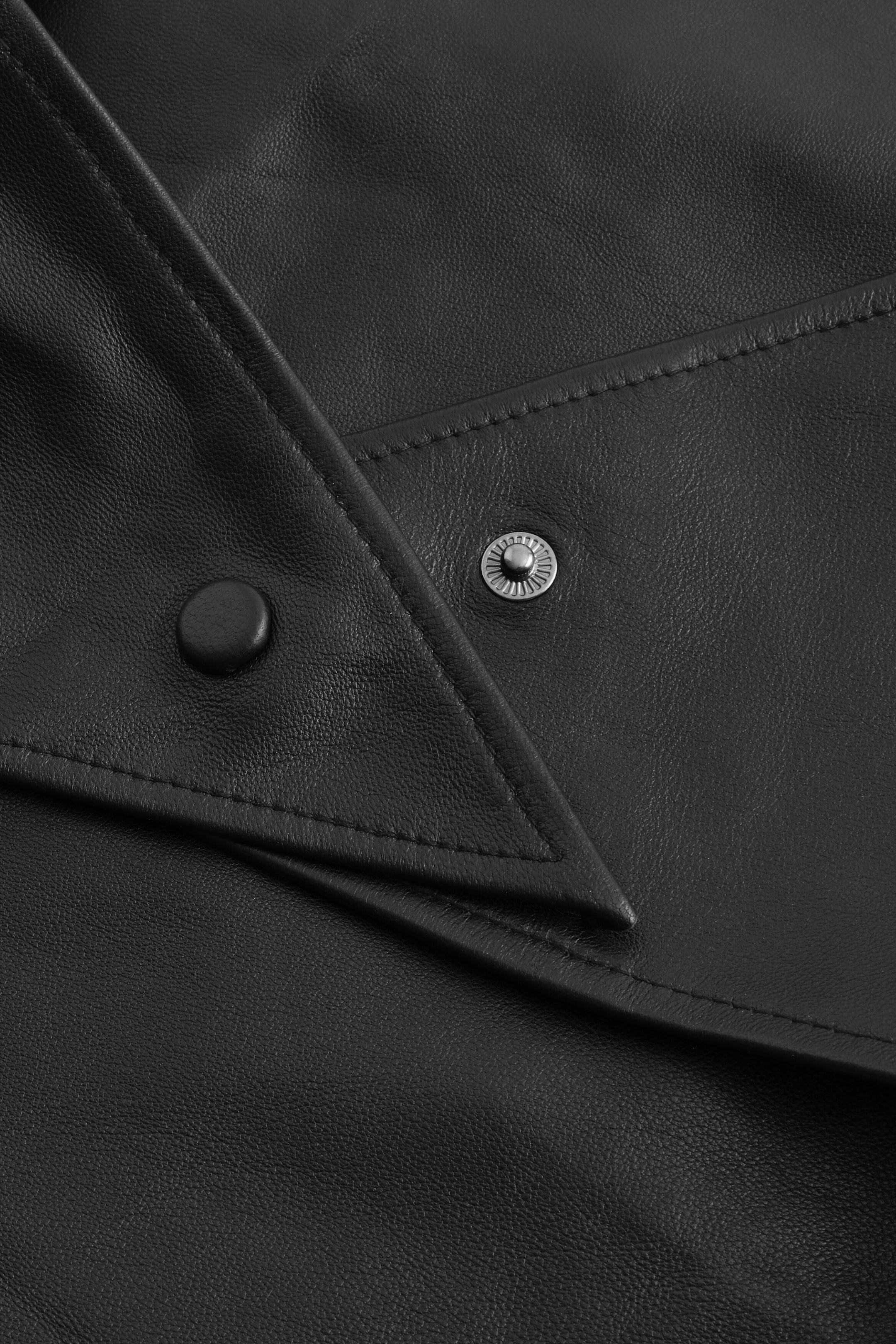 The black leather hood is showing a close-up detail of its snap-button fastening mechanism, featuring a pointed leather flap with a covered snap and an exposed metal snap socket set against smooth, topstitched leather. The black leather hood is showing a close-up detail of its snap-button fastening mechanism, featuring a pointed leather flap with a covered snap and an exposed metal snap socket set against smooth, topstitched leather.