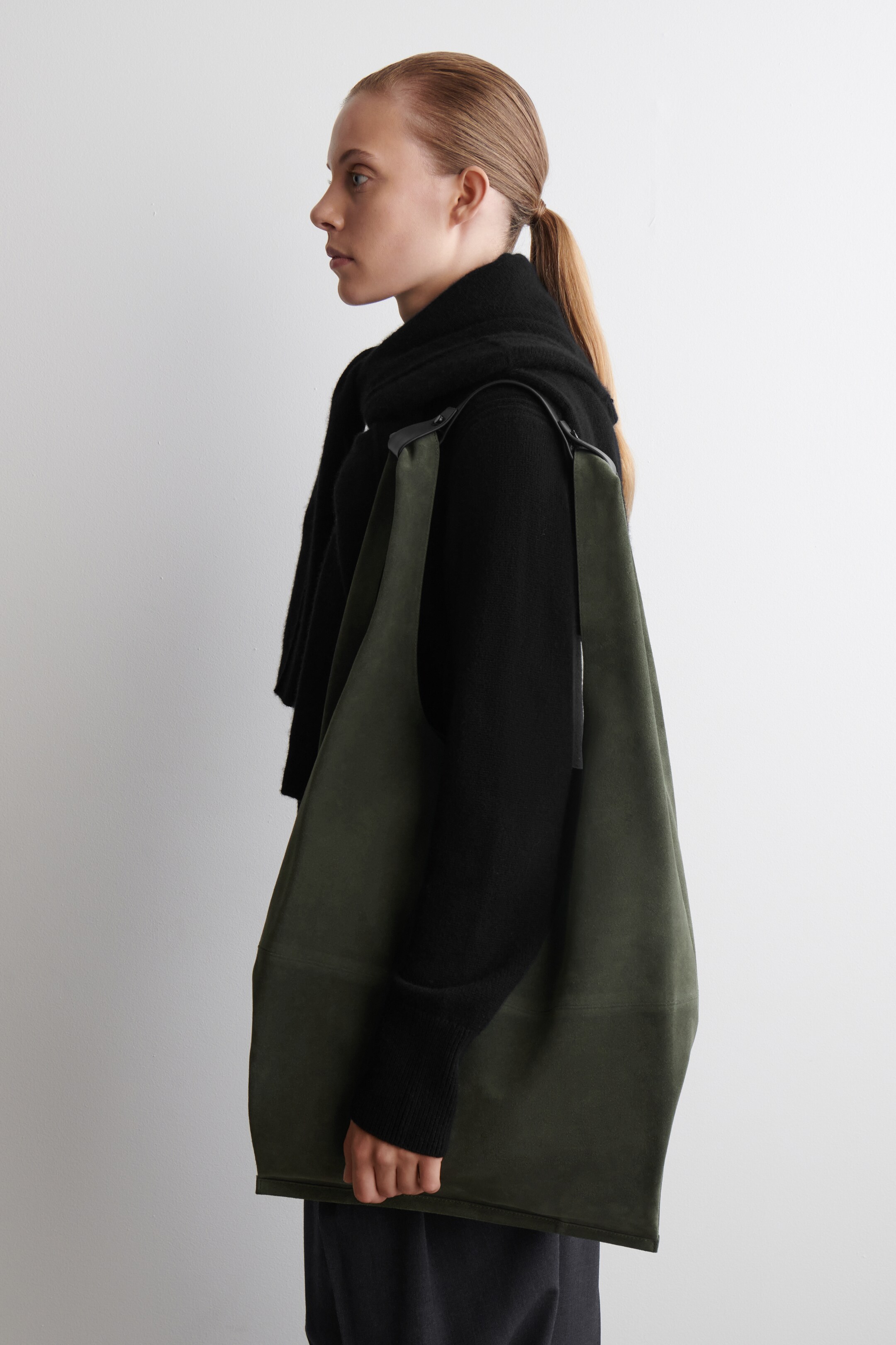 Agrandir l'image: Un sac à bandoulière vert olive, à structure souple, est porté sur le côté droit du porteur, orienté vers la droite du spectateur et pend librement. Une large sangle noire s'étend depuis ses coins supérieurs, et une couture horizontale est visible sur sa partie inférieure.