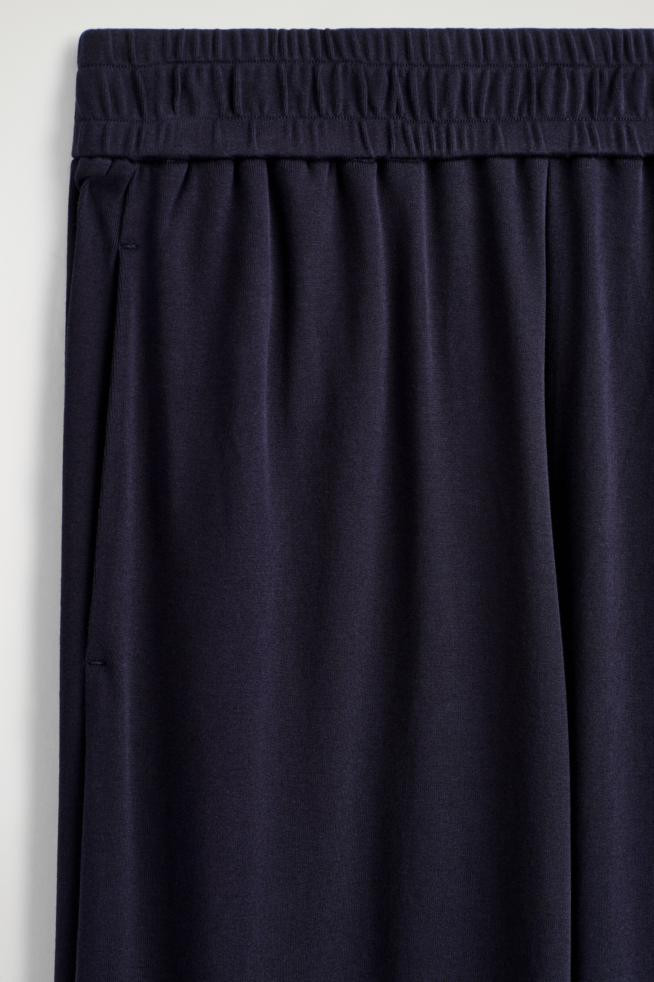 Agrandir l'image: Les Joggers en Jersey Fluide présentent un tissu en jersey fluide bleu foncé à l'avant, avec une large ceinture élastiquée froncée et une poche discrète dans la couture latérale gauche.