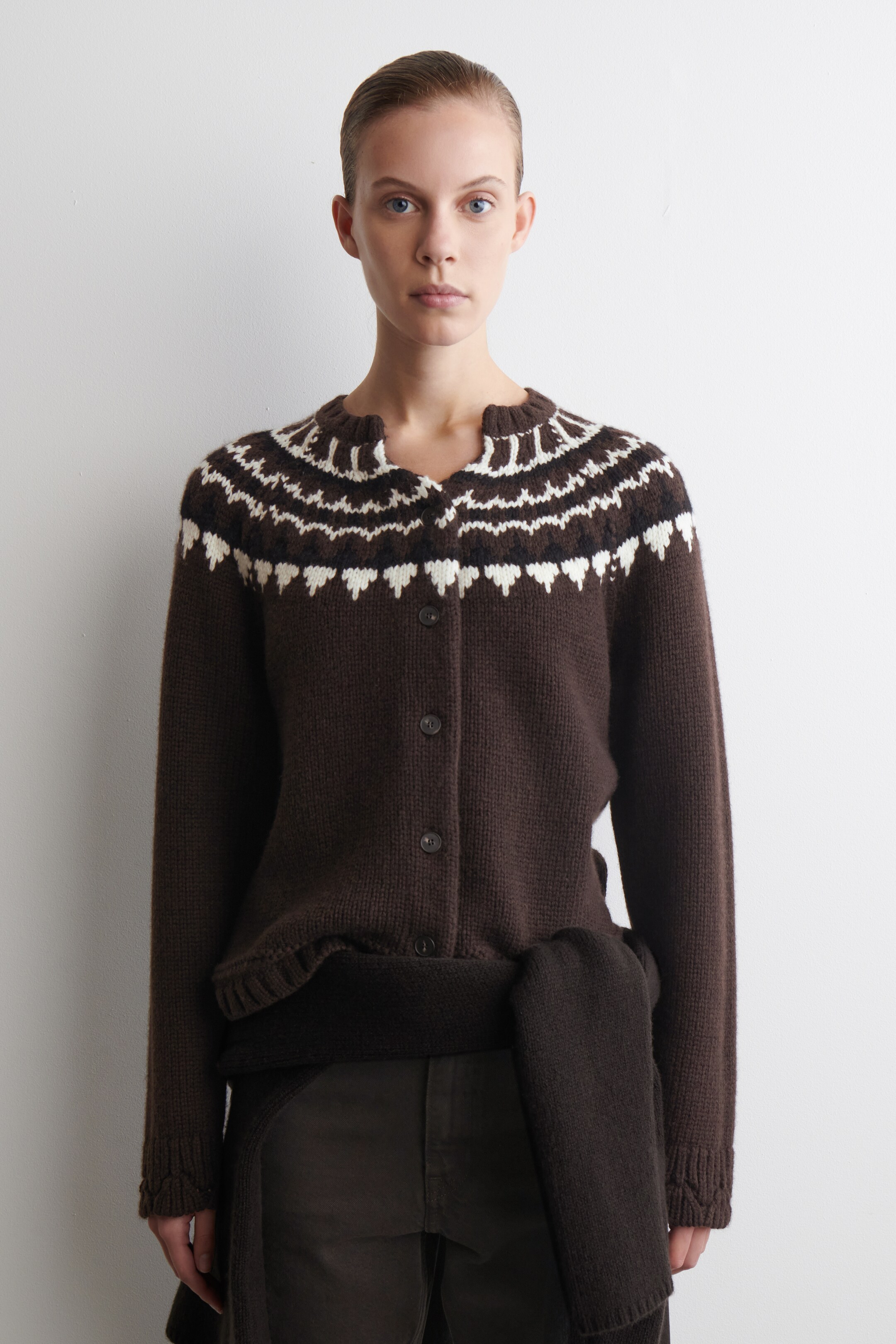 Visualizza immagine più grande: Un cardigan aderente marrone scuro viene presentato di FRONTE, mostrando un motivo fairisle in bianco e marrone scuro sul petto e sulle spalle. Presenta una chiusura frontale con bottoni, polsini e orlo a coste e uno scollo a coste smerlate. Un indumento marrone scuro è legato intorno alla vita.