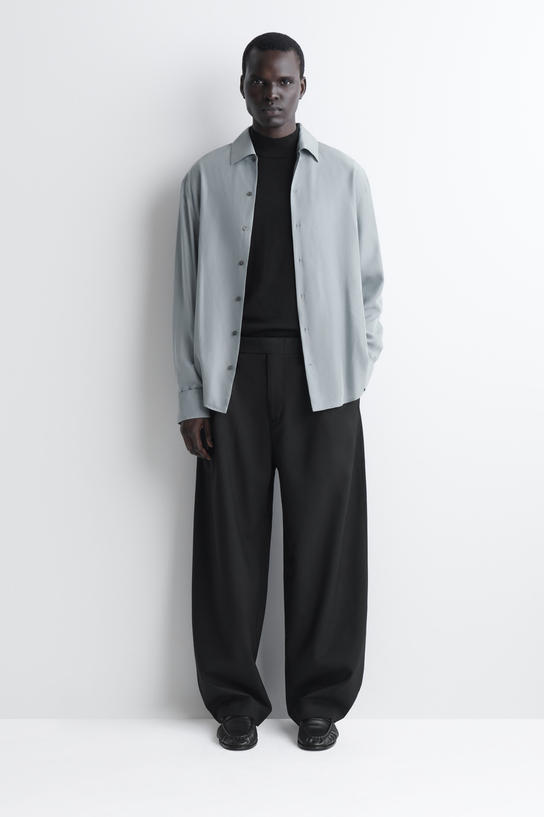 Diese schwarzen Barrel-Leg-Hosen aus Wolle mit Relaxed-Fit werden von vorne gezeigt und verfügen über ein hoch tailliertes, weit geschnittenes Design, das sich leicht nach außen wölbt und am Knöchel schmaler zuläuft. Die Hose ist mit einem in den Bund gesteckten schwarzen Rollkragenpullover und einem offen getragenen hellblauen Hemd gestylt, wodurch der klare, strukturierte Bund und der entspannte Fall hervorgehoben werden. Diese schwarzen Barrel-Leg-Hosen aus Wolle mit Relaxed-Fit werden von vorne gezeigt und verfügen über ein hoch tailliertes, weit geschnittenes Design, das sich leicht nach außen wölbt und am Knöchel schmaler zuläuft. Die Hose ist mit einem in den Bund gesteckten schwarzen Rollkragenpullover und einem offen getragenen hellblauen Hemd gestylt, wodurch der klare, strukturierte Bund und der entspannte Fall hervorgehoben werden.