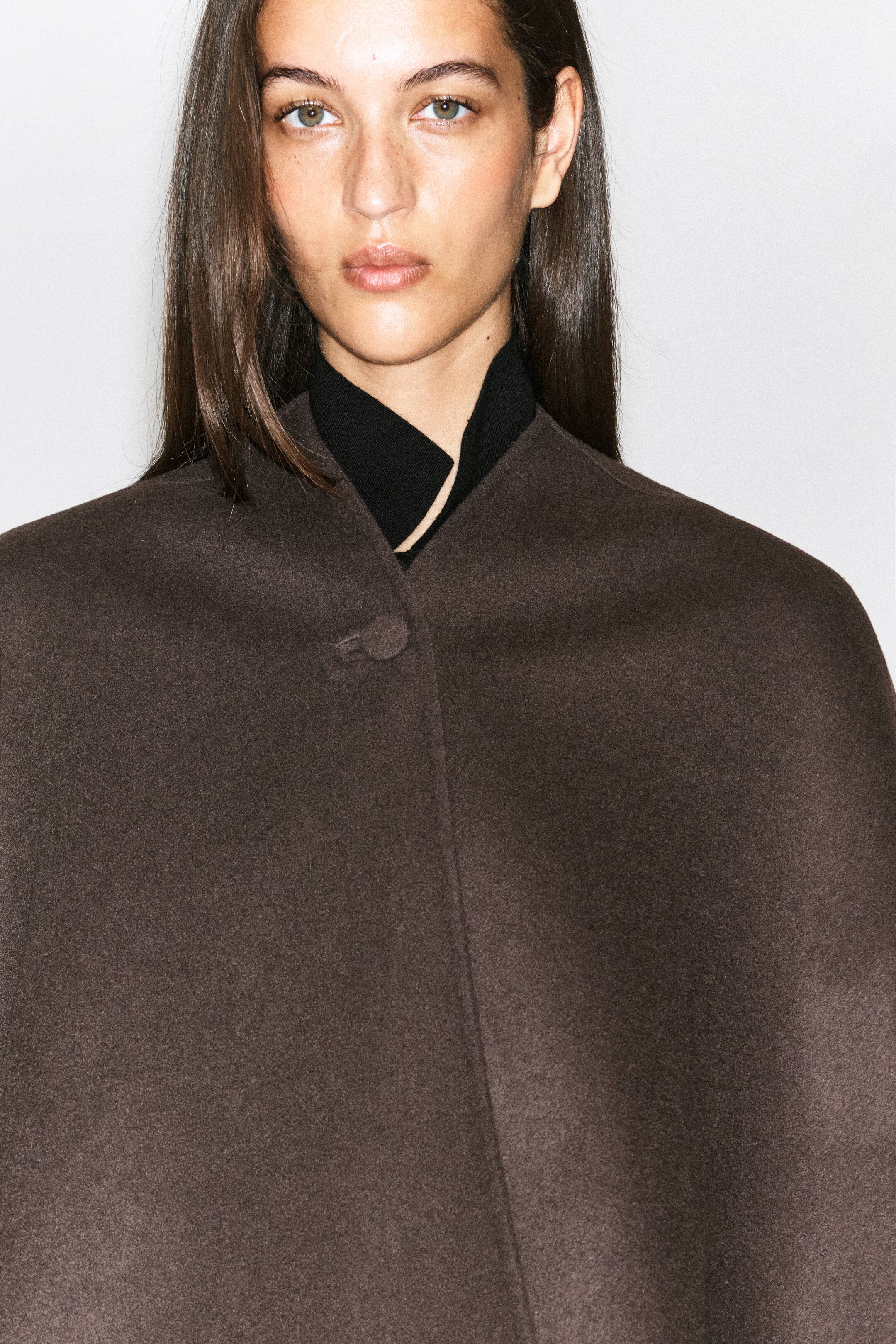 Agrandir l'image: Une cape en mélange de laine double face, de couleur marron foncé, est portée par le mannequin par-dessus un vêtement noir à col montant. La cape présente un col sans col, fermé par un seul bouton en haut.