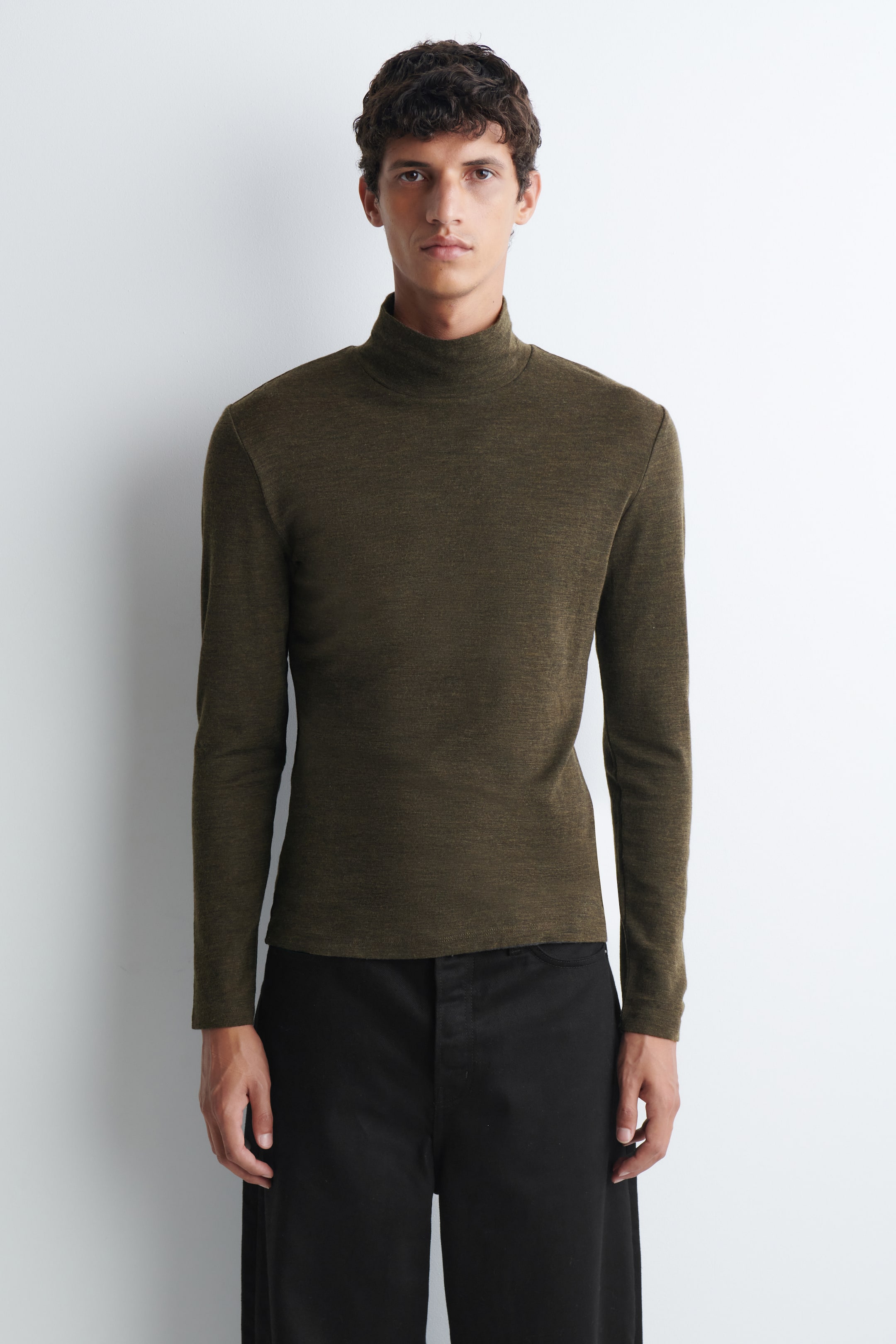 Visualizza l'immagine ingrandita: Un top slim-fit in jersey di lana con collo alto color verde oliva è indossato dalla modella, mettendo in risalto il suo collo alto, le maniche lunghe e la trama fine. Il top è indossato senza essere rimboccato e abbinato a pantaloni neri.