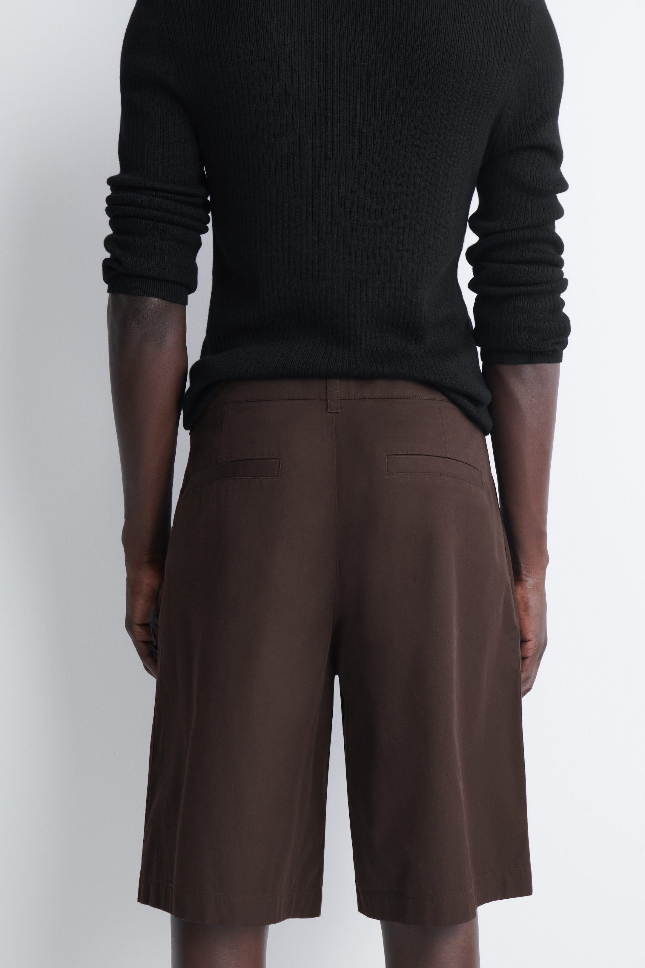 Tailored pleated cotton shorts in dark brown are shown from the back, featuring a relaxed fit, belt loops, and two rear welt pockets. The shorts are styled with a black ribbed top that sits just above the waistband. Tailored pleated cotton shorts in dark brown are shown from the back, featuring a relaxed fit, belt loops, and two rear welt pockets. The shorts are styled with a black ribbed top that sits just above the waistband.
